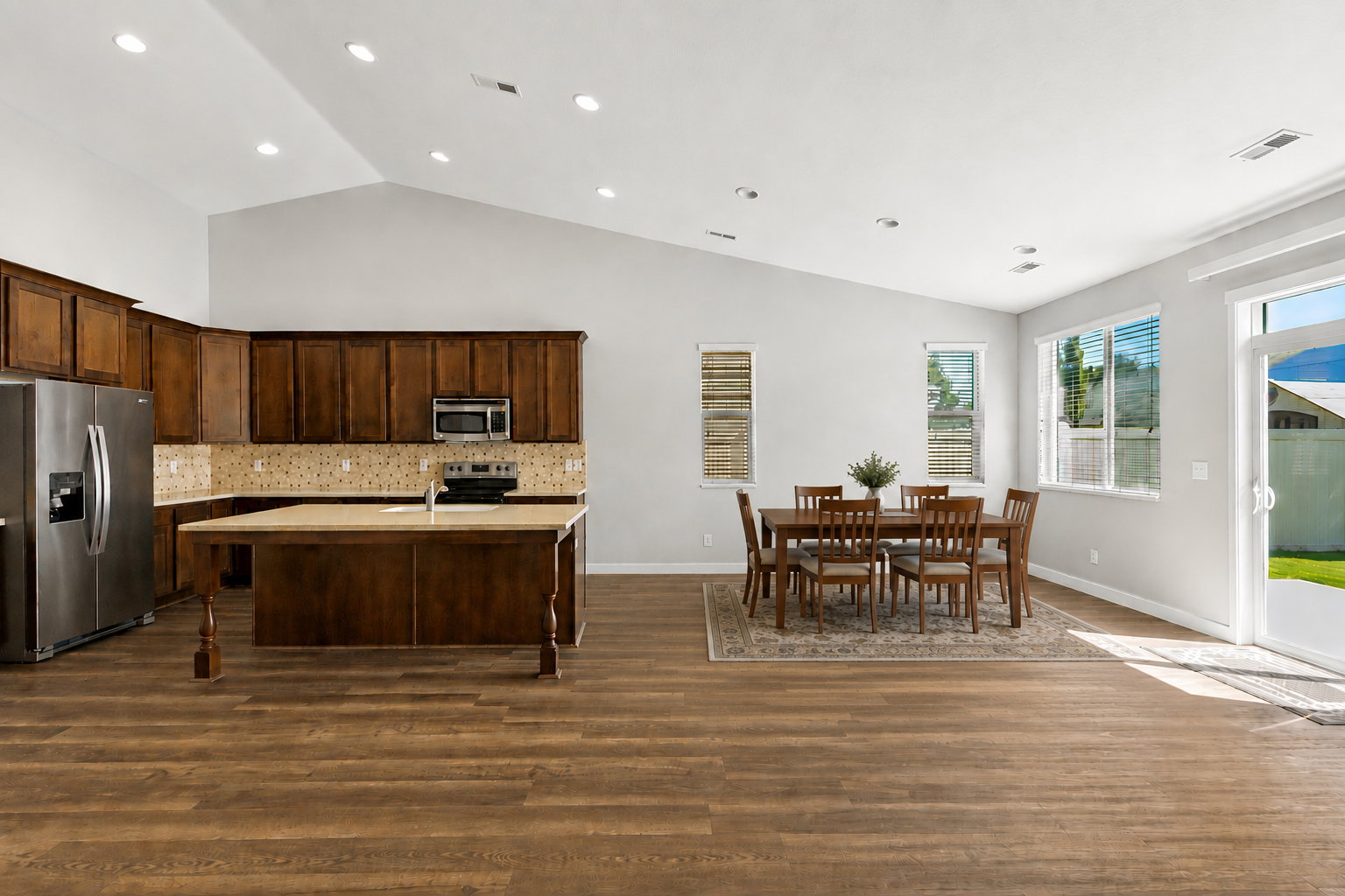 AI staged table in open-concept living area featuring wood-finish flooring, a kitchen island with built-in sink, stainless steel appliances, and abundant natural light from multiple windows and a glass patio door