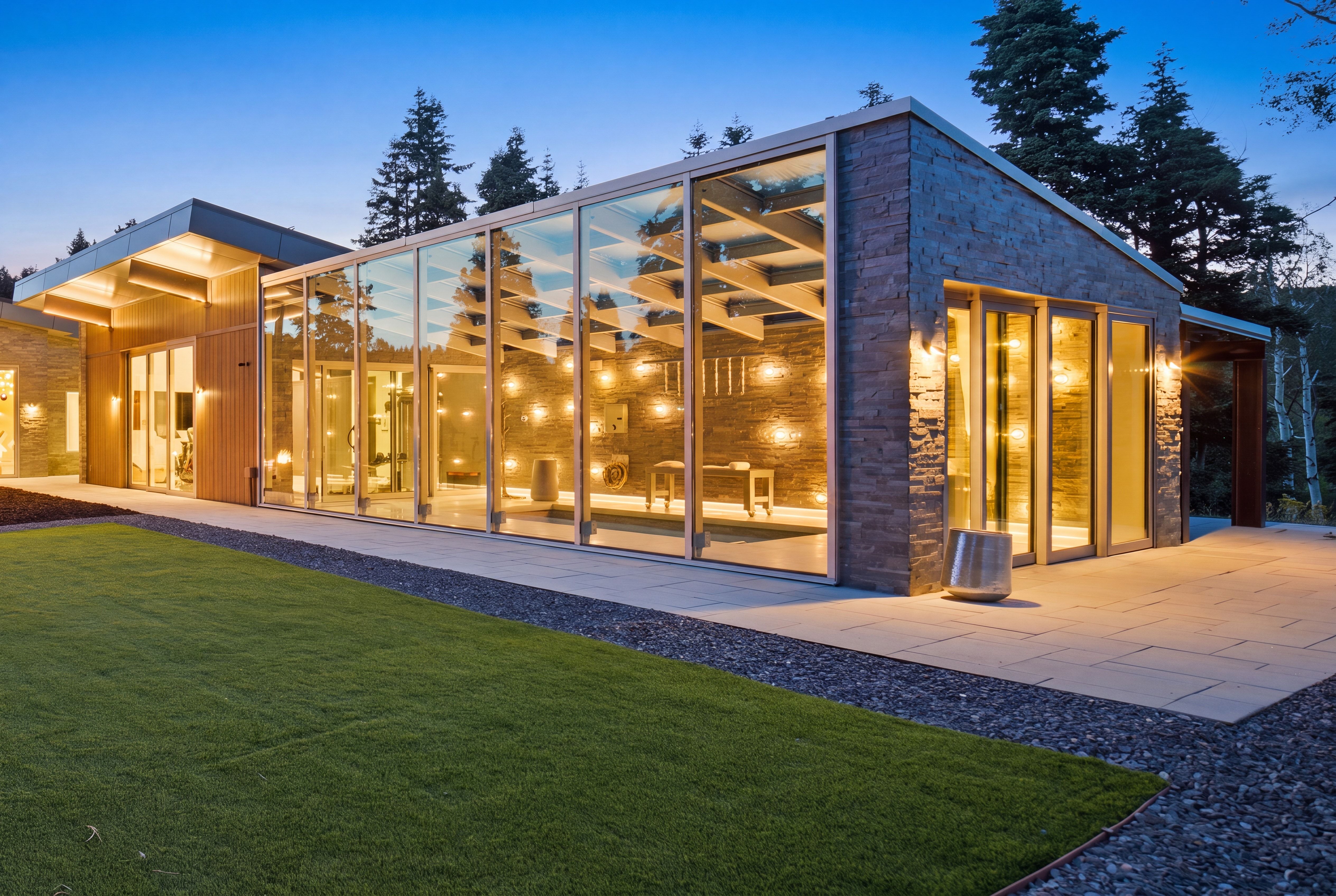 Modern architectural design featuring extensive glass paneling, natural stone accents, wood-finish siding, a large patio area, and a manicured lawn
