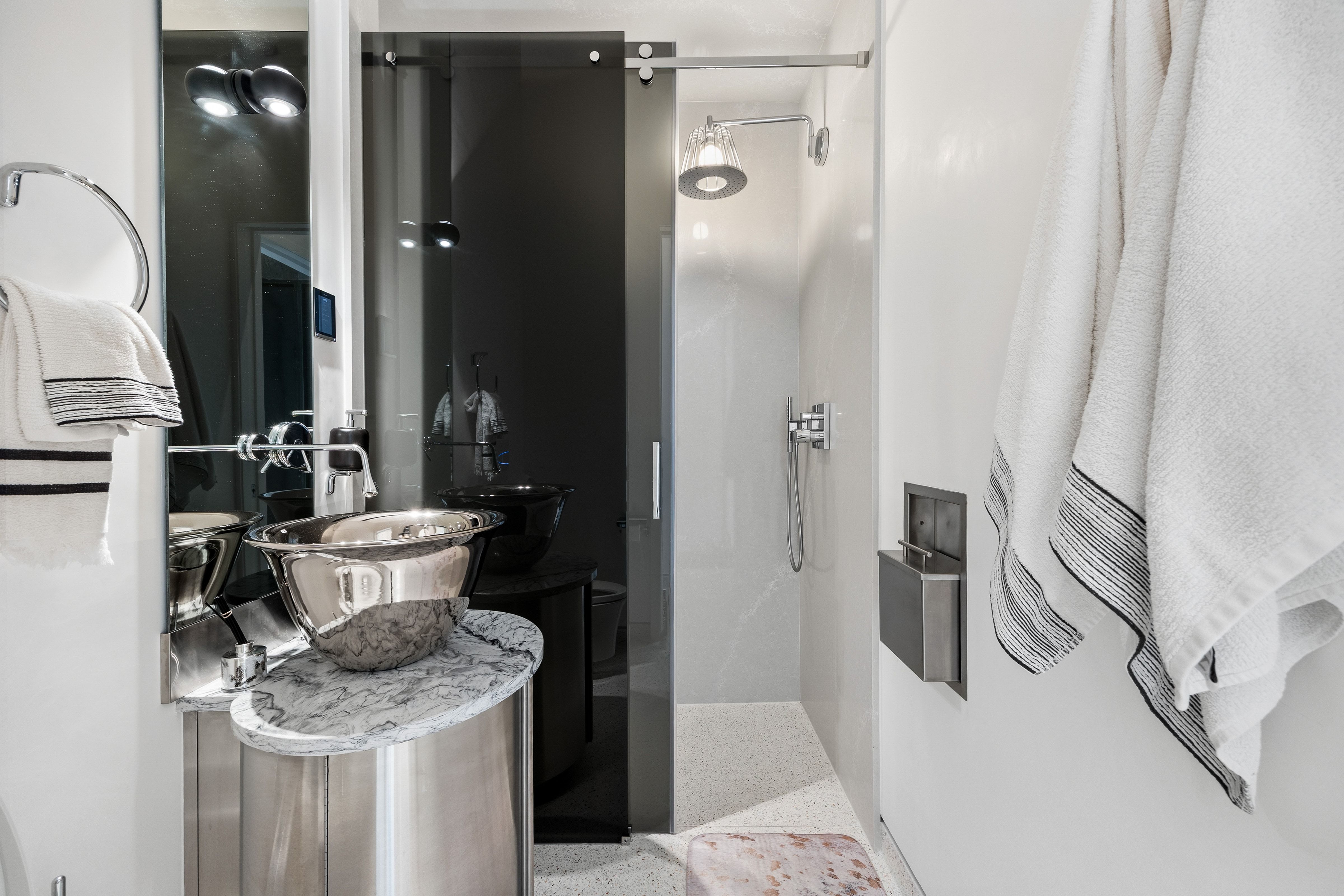 Modern bathroom featuring a glass-enclosed shower with a rainfall showerhead, a metal vessel sink, a stone-finish countertop, and built-in wall storage