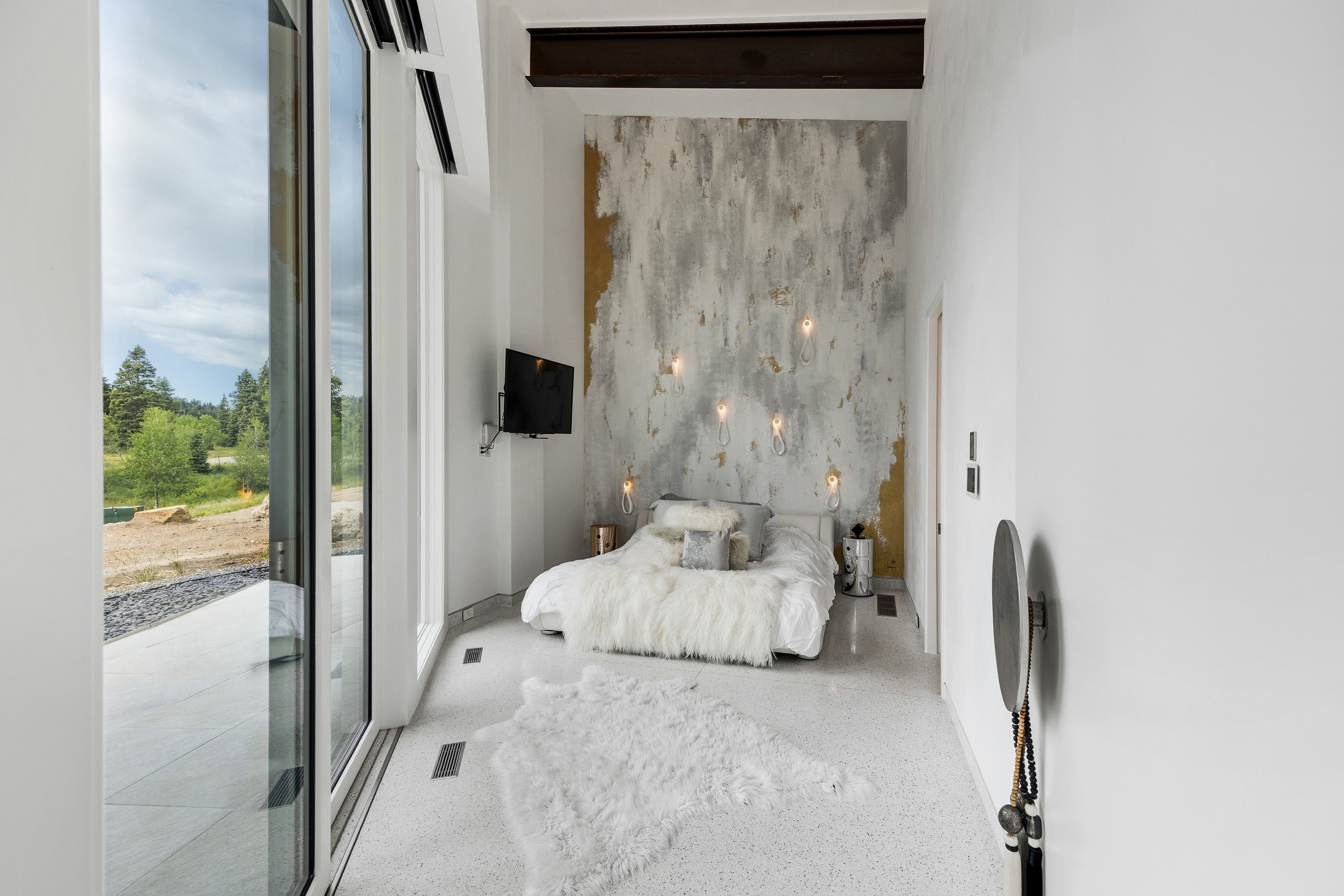 Spacious room featuring a full wall of floor-to-ceiling windows, a textured accent wall with wall-mounted light fixtures, a mounted flat-screen television, and light-toned terrazzo flooring