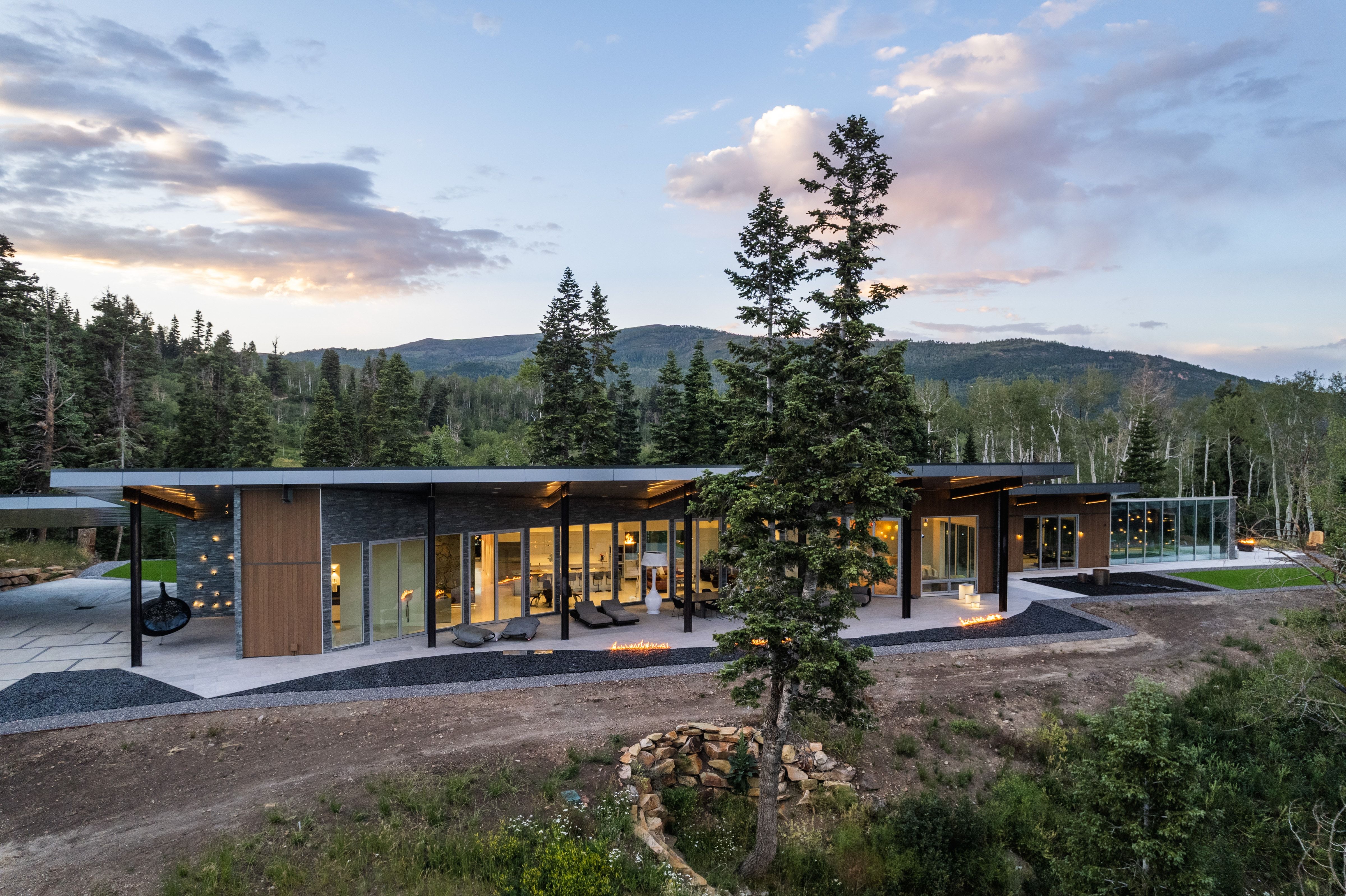 Contemporary exterior with a flat roofline, extensive glass doors, stone and wood-panel accents, a spacious stone patio, and integrated outdoor lighting