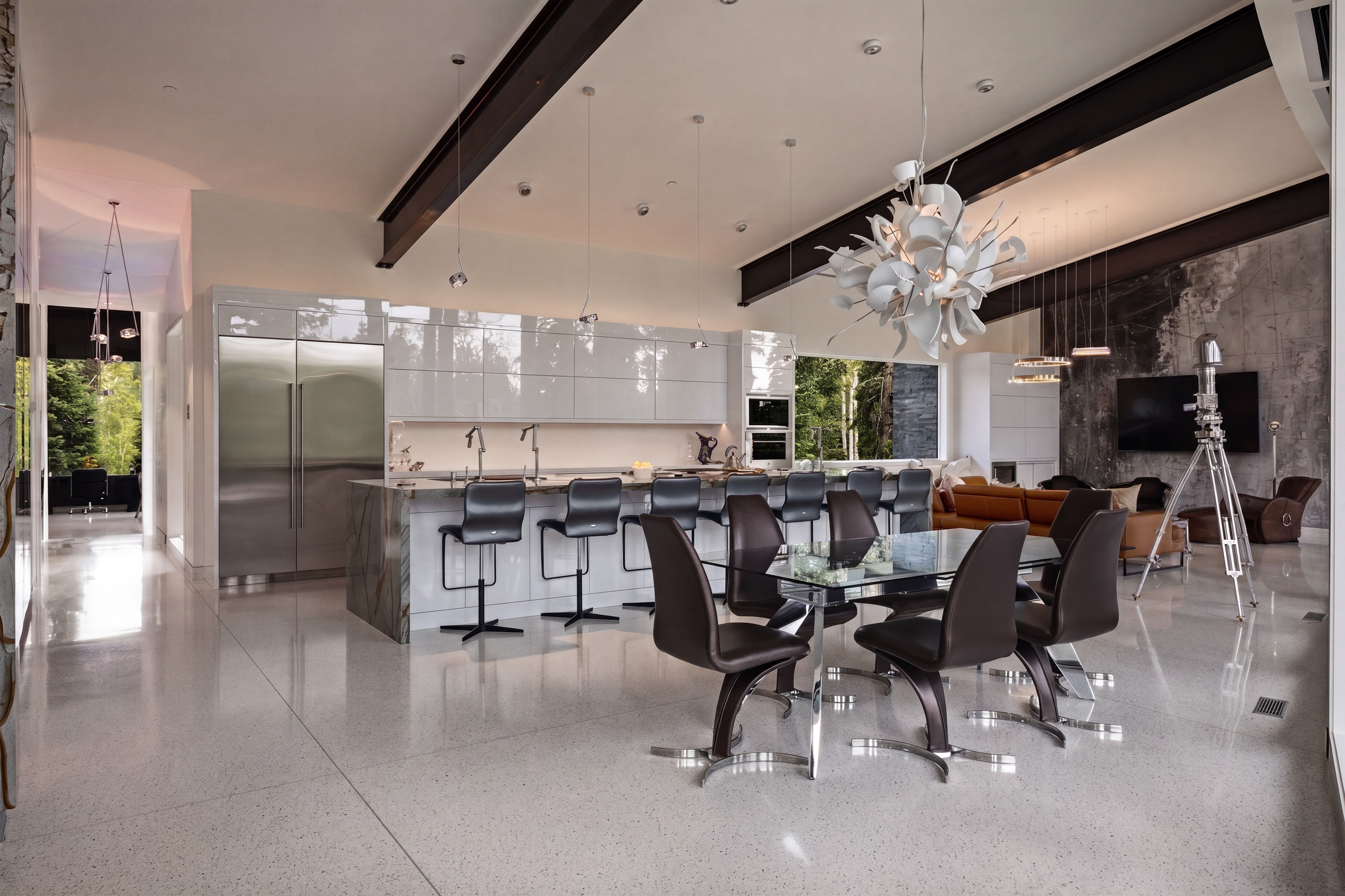 Open-concept living space featuring high vaulted ceilings with exposed beams, polished terrazzo flooring, a modern kitchen with high-gloss cabinetry and integrated stainless steel appliances, a large center island with bar seating, and a unique sculptural