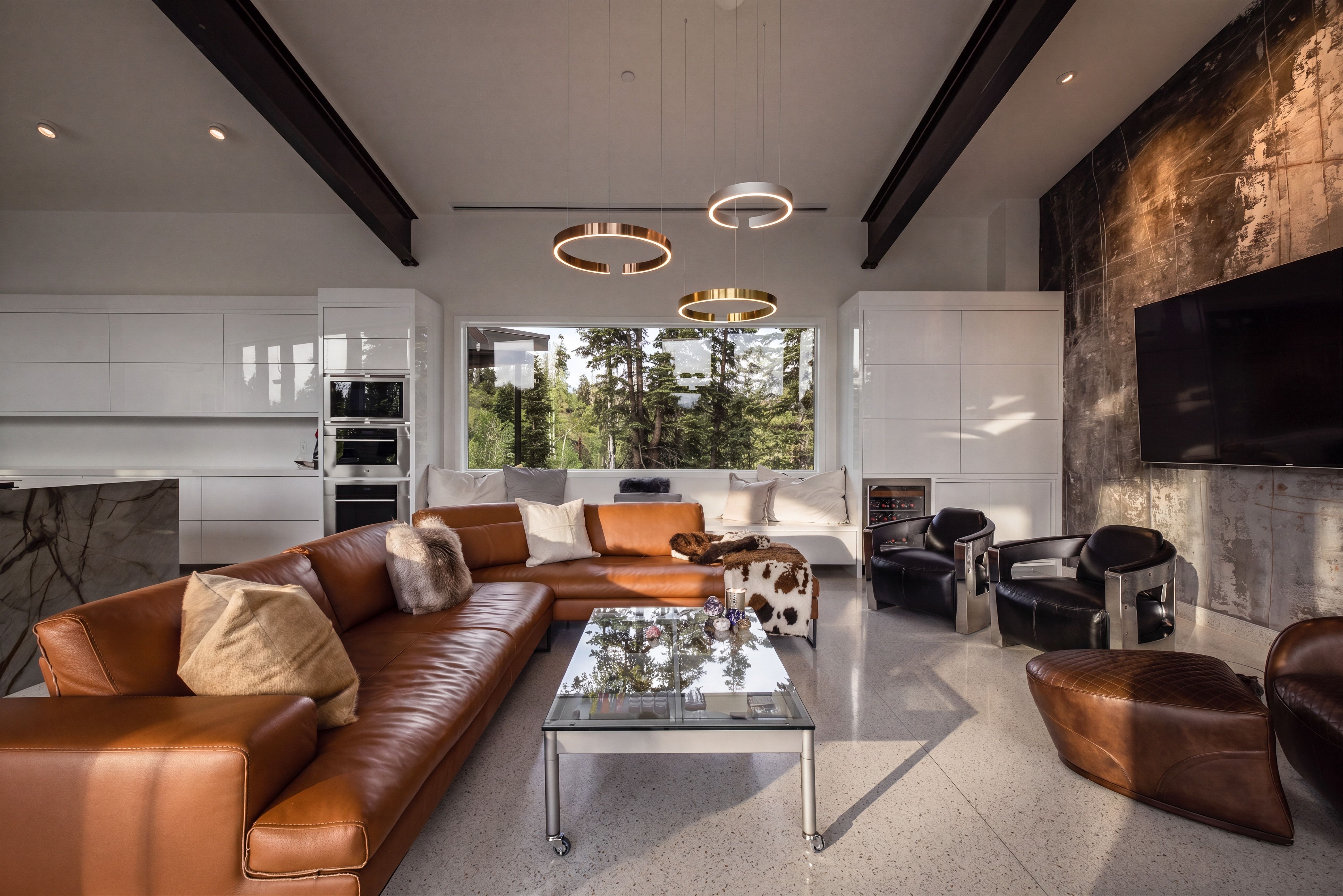 Open-concept living space featuring high ceilings with exposed beams, an integrated kitchen with built-in appliances and white flat-panel cabinetry, a large picture window, and stone flooring