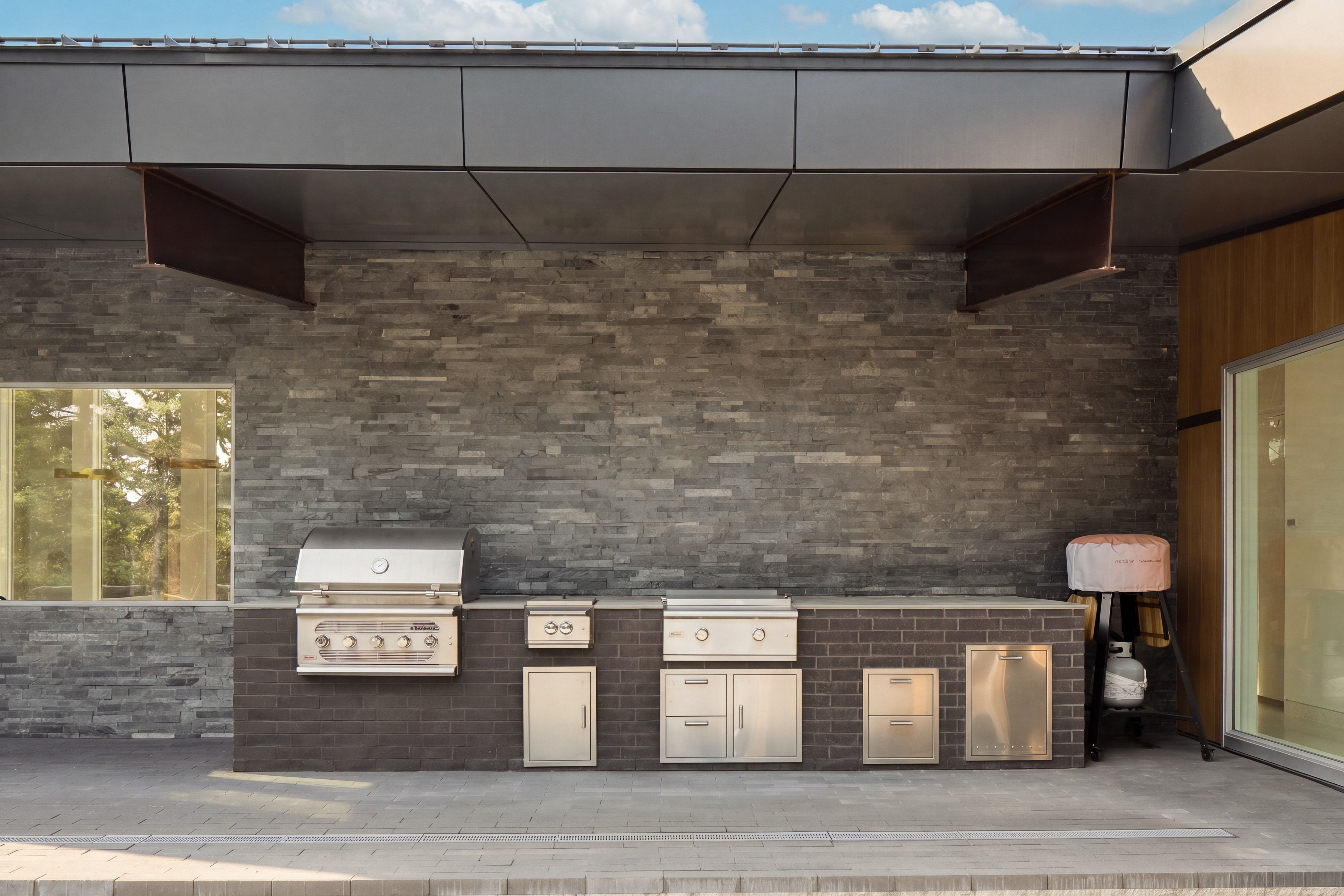 Outdoor kitchen featuring integrated stainless steel appliances, brick base, stacked stone wall, and a covered patio area
