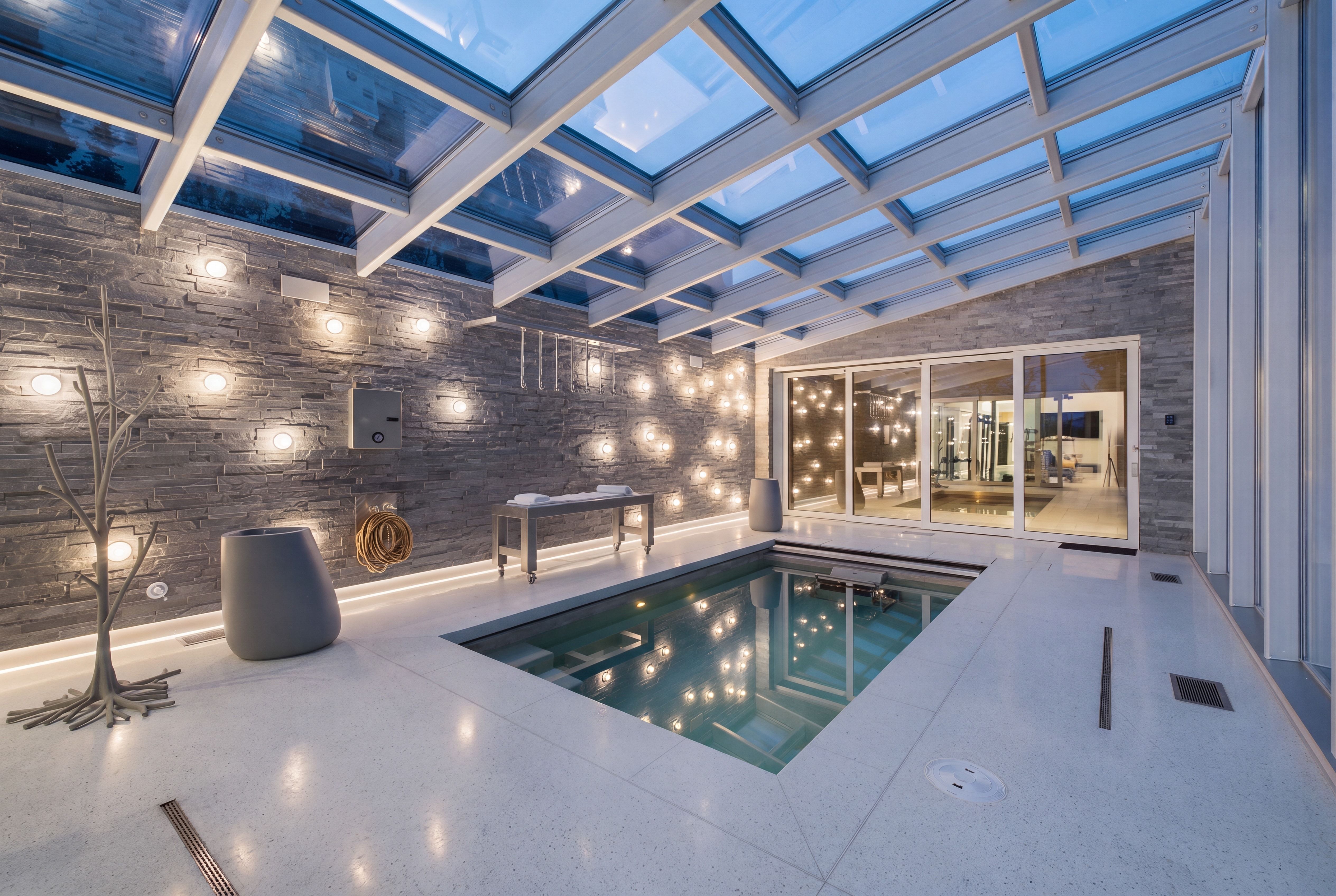 Indoor pool area featuring a glass-paneled ceiling, stone accent wall with multiple wall sconces, large format tile flooring, and sliding glass doors to an interior space