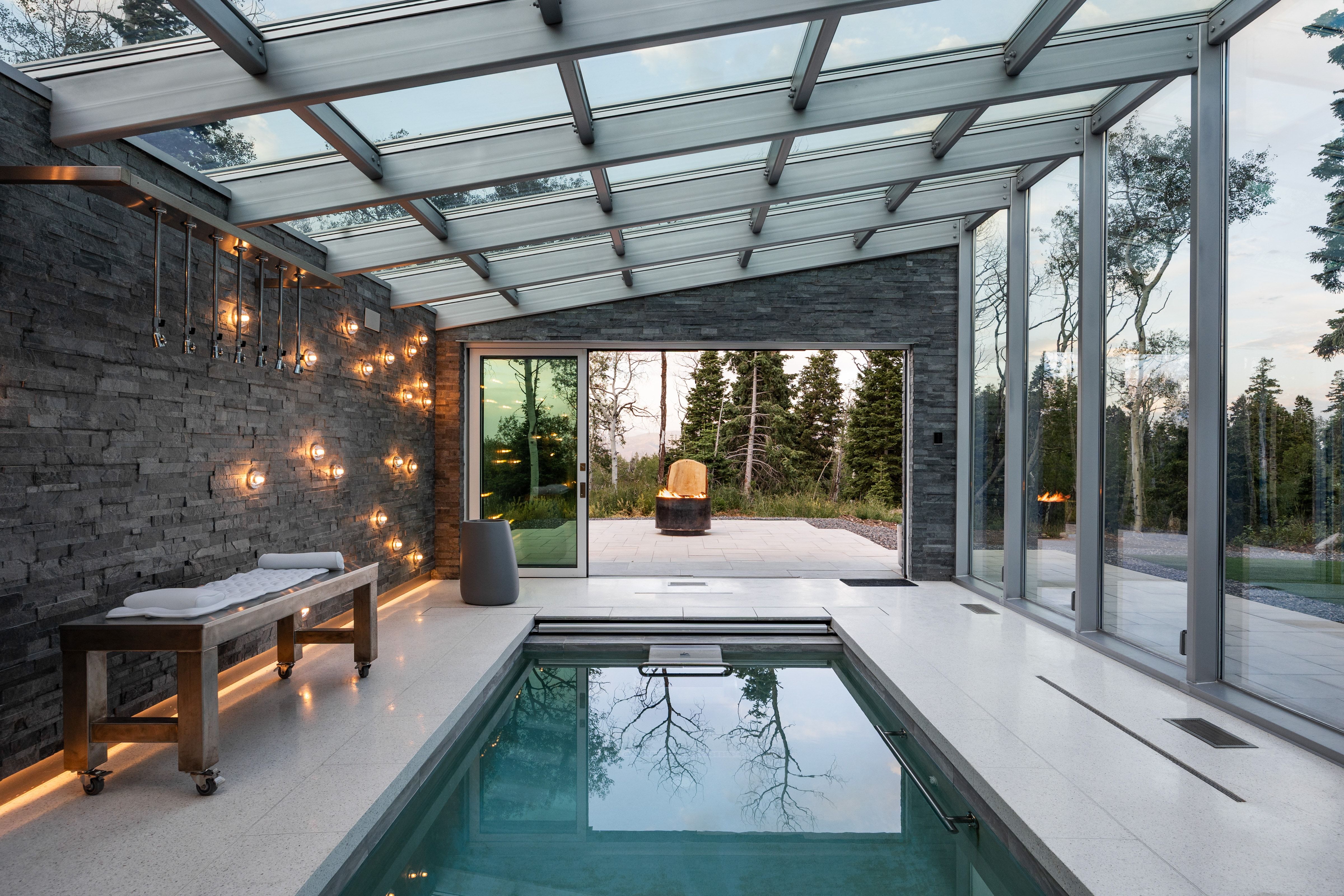 Indoor swimming pool area featuring a glass ceiling, stone accent wall, large format floor tiles, and full-height glass panels opening to an outdoor patio