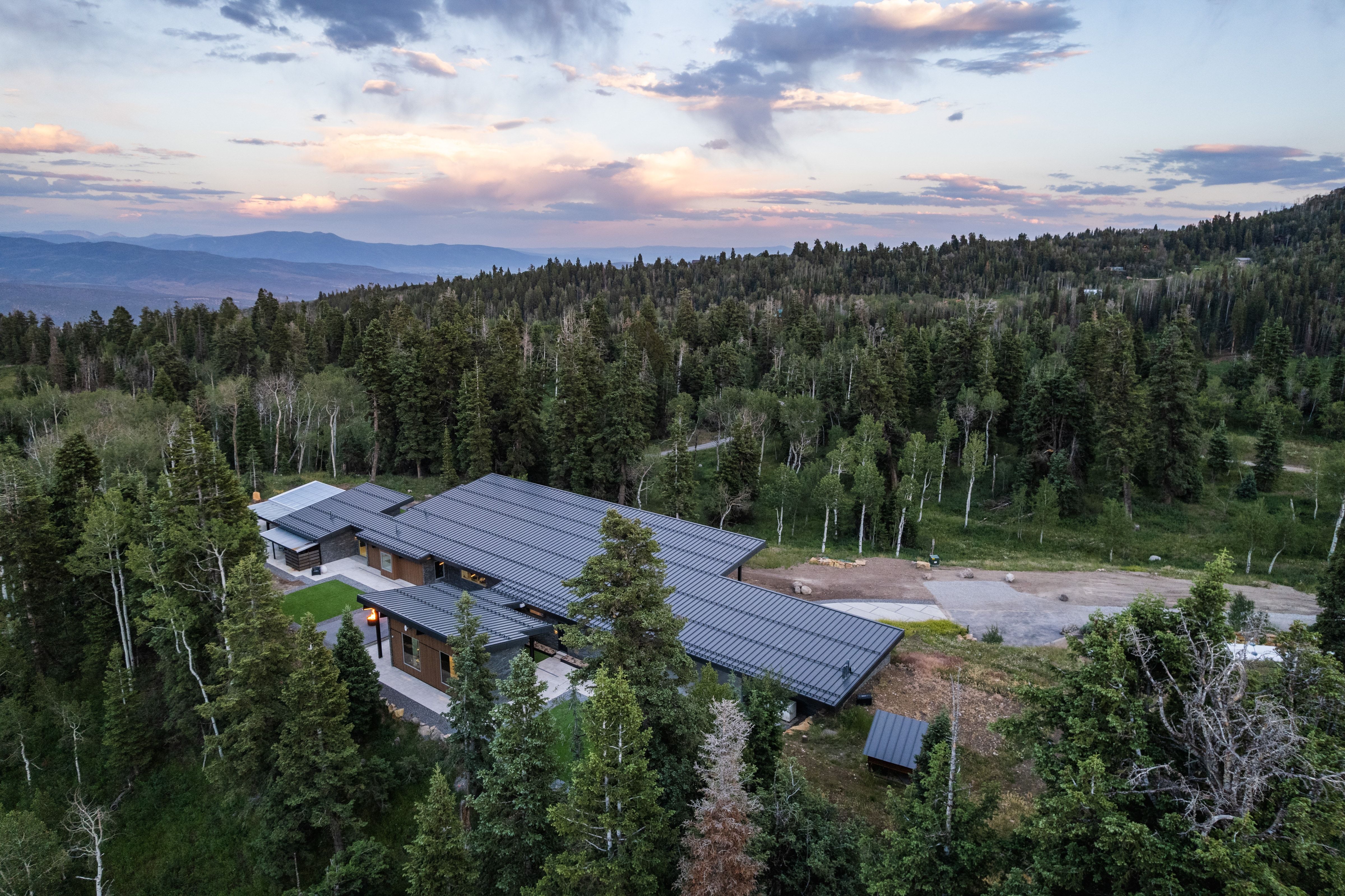 Expansive property featuring a modern architectural design, multiple metal roofs, and a surrounding forest of mature trees