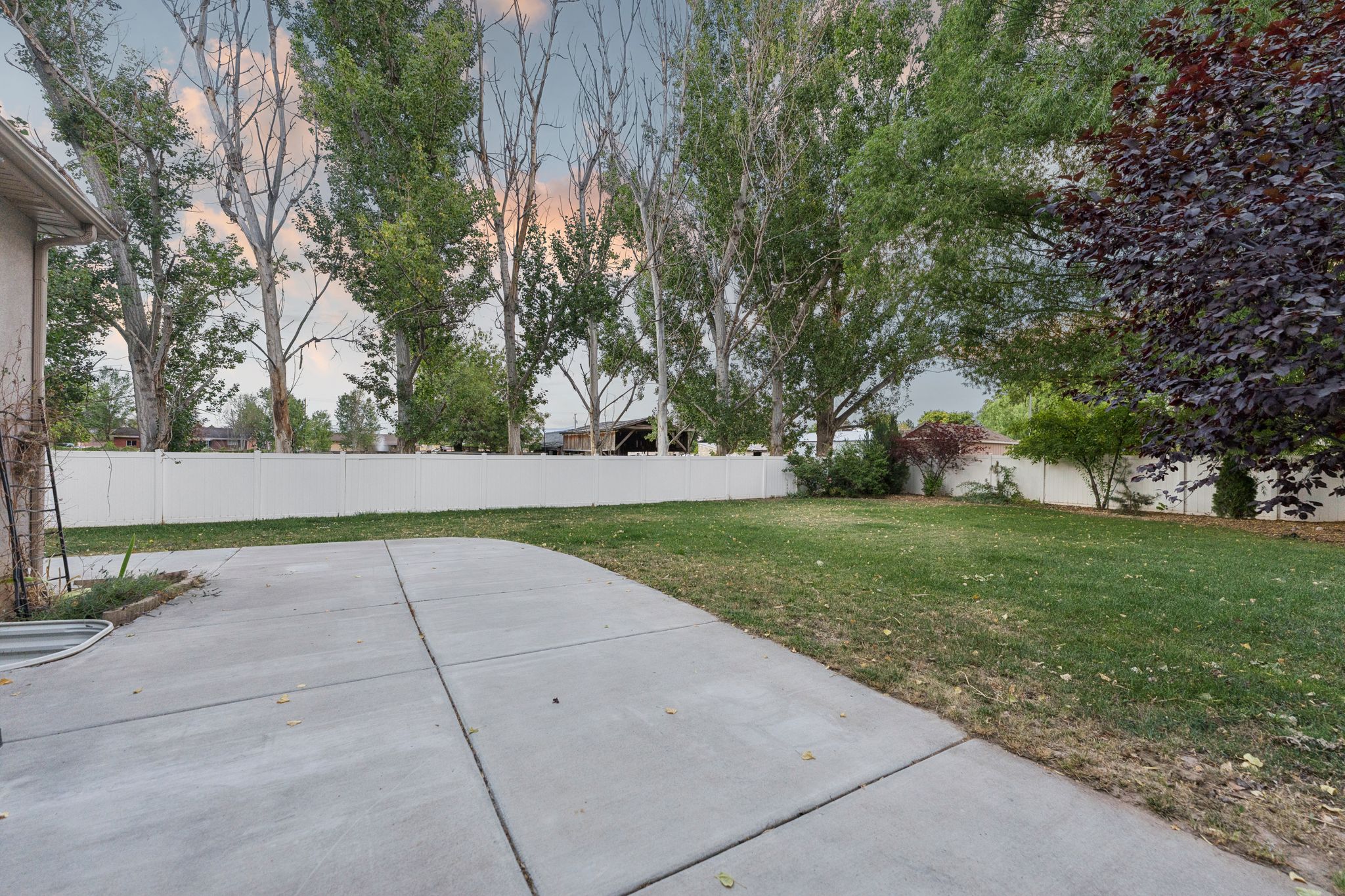 Expansive lawn area featuring a white privacy fence, mature trees, concrete patio space, and varied landscaping