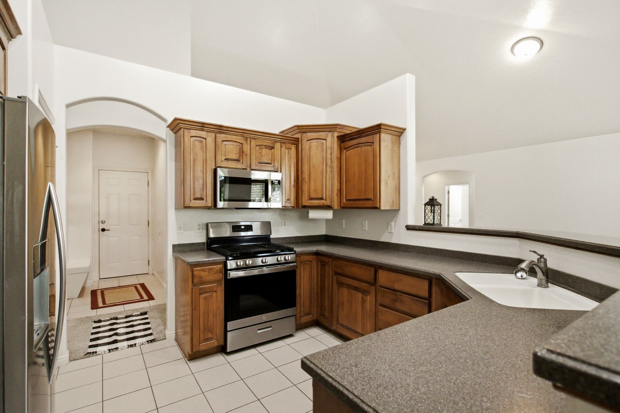 Kitchen featuring wood cabinetry, stainless steel appliances, a corner sink with a gooseneck faucet, composite countertops, and white tile flooring