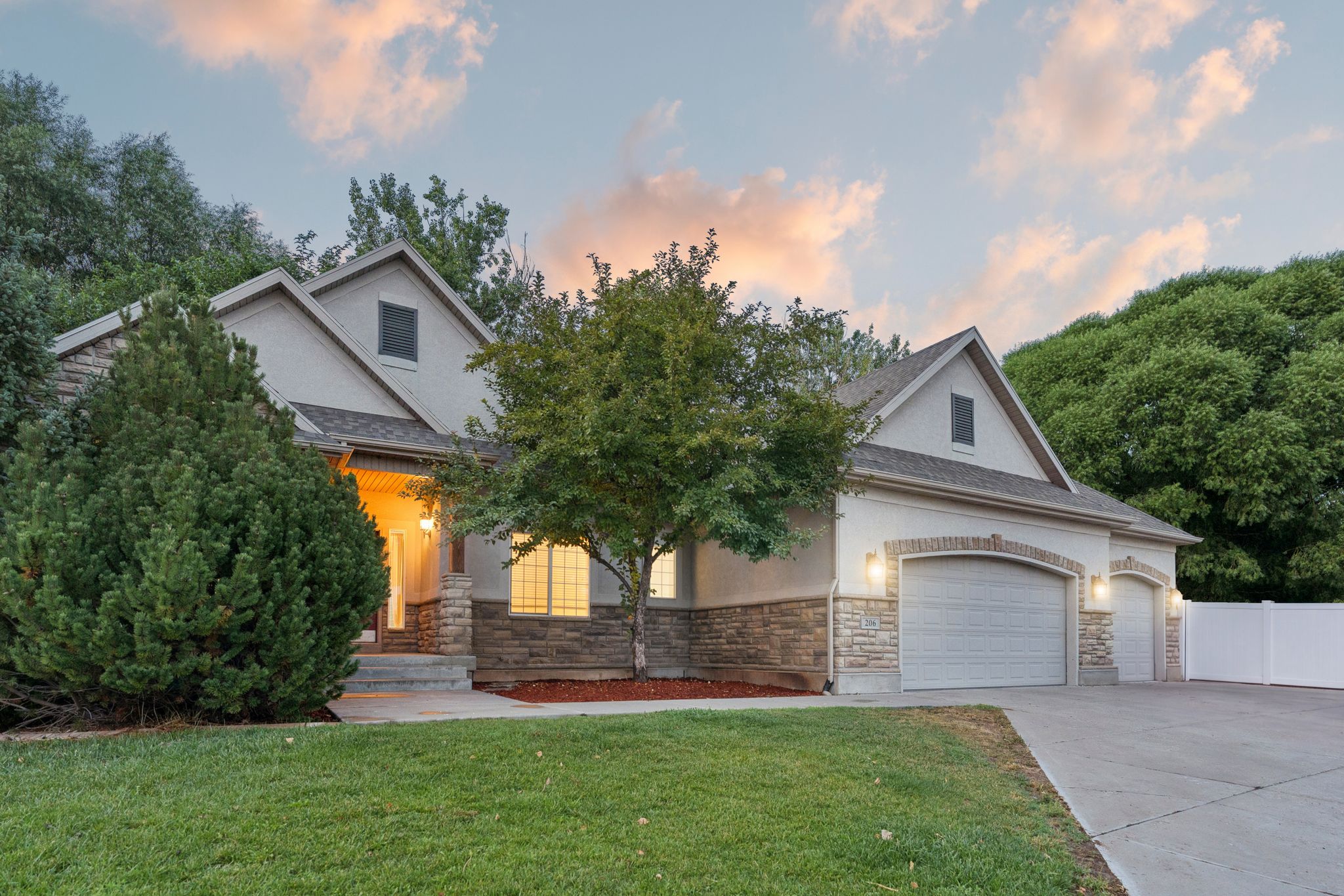 Two-story stucco and stone-faced residence featuring a two-car garage with arched brick detailing, mature landscaping, and a concrete driveway