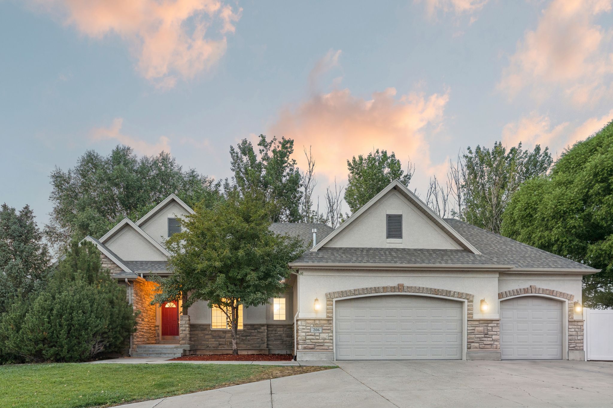 Exterior showcasing a stucco and stone facade, a red entry door, a two-car garage, an additional single-car garage, and mature landscaping