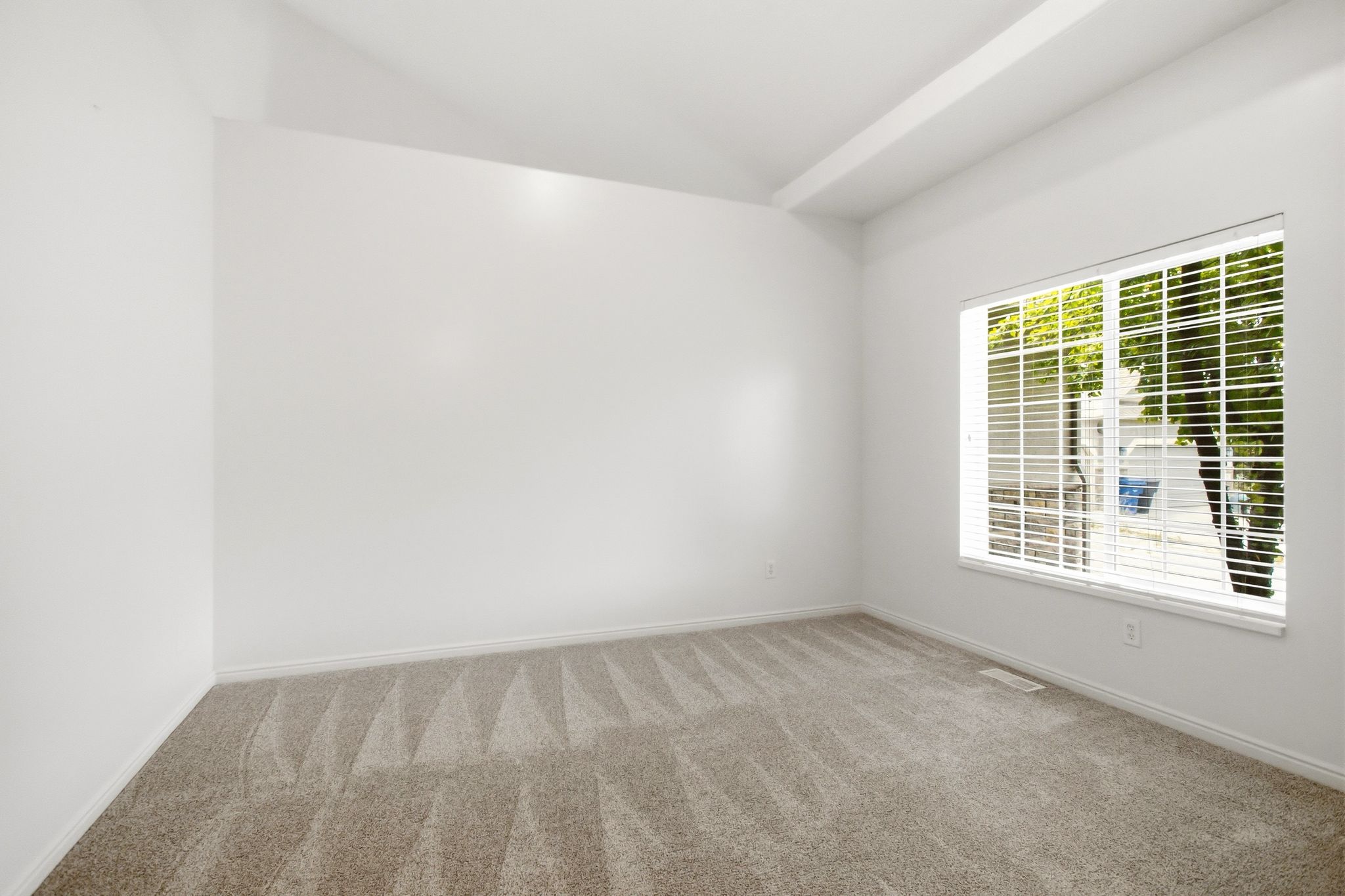 Bright interior space featuring a large window with horizontal blinds, neutral-toned carpeting, white baseboards, and a vaulted ceiling