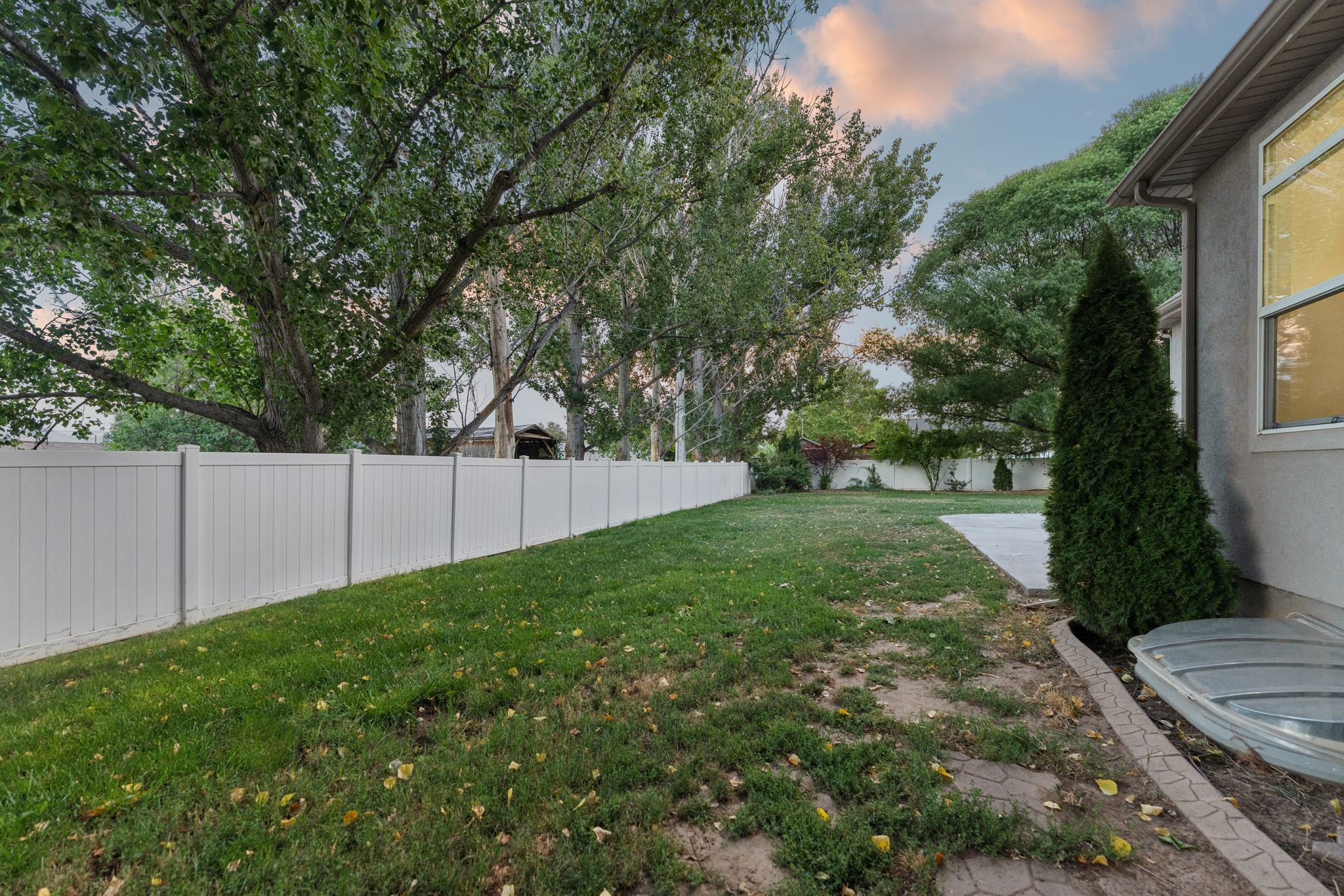 Expansive backyard featuring a white vinyl privacy fence, mature trees, a concrete patio area, and a stucco exterior