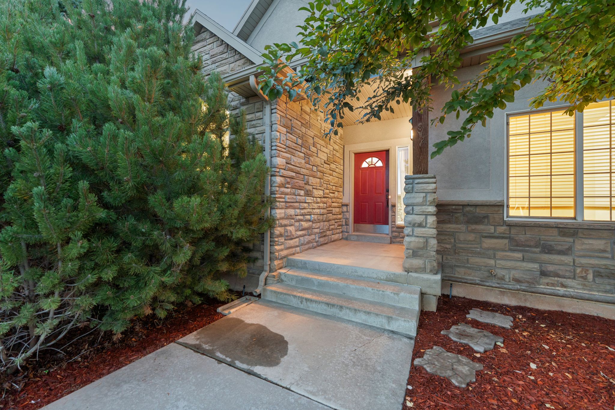 Welcoming entry with a red panel door featuring an arched glass insert, stone veneer siding, concrete steps, and integrated outdoor lighting