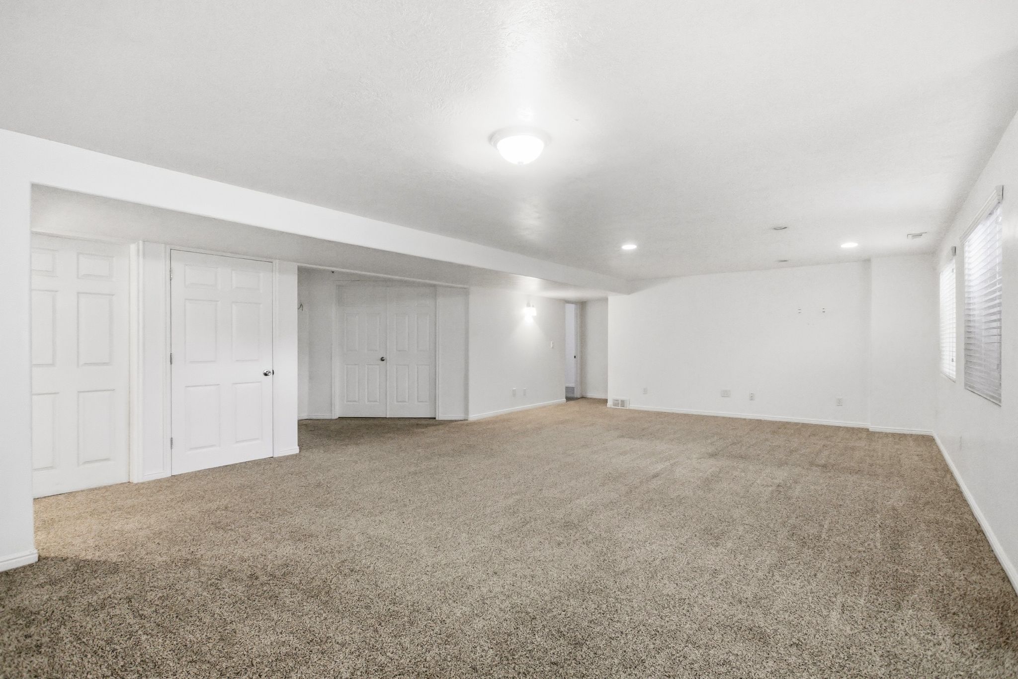 Spacious open room featuring neutral-toned carpet flooring, white painted walls, multiple white panel doors, recessed lighting, and windows with blinds
