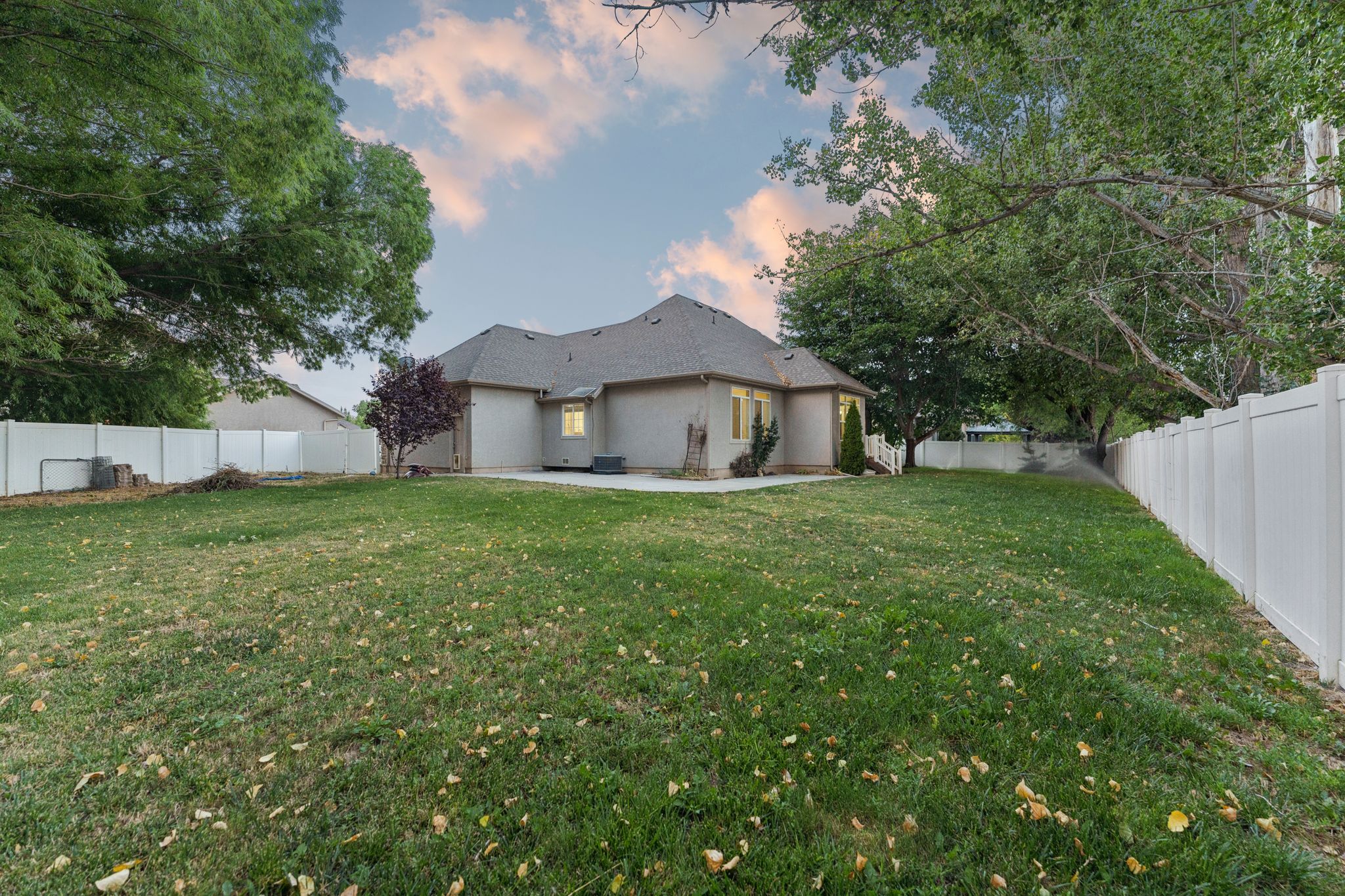 Expansive backyard featuring a large grass lawn, mature trees, a white privacy fence, and a concrete patio
