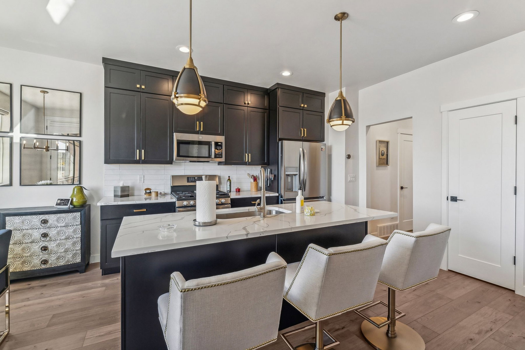 Open-concept kitchen featuring dark cabinetry with gold-toned hardware, stainless steel appliances, a subway tile backsplash, and a large island with a light-toned countertop