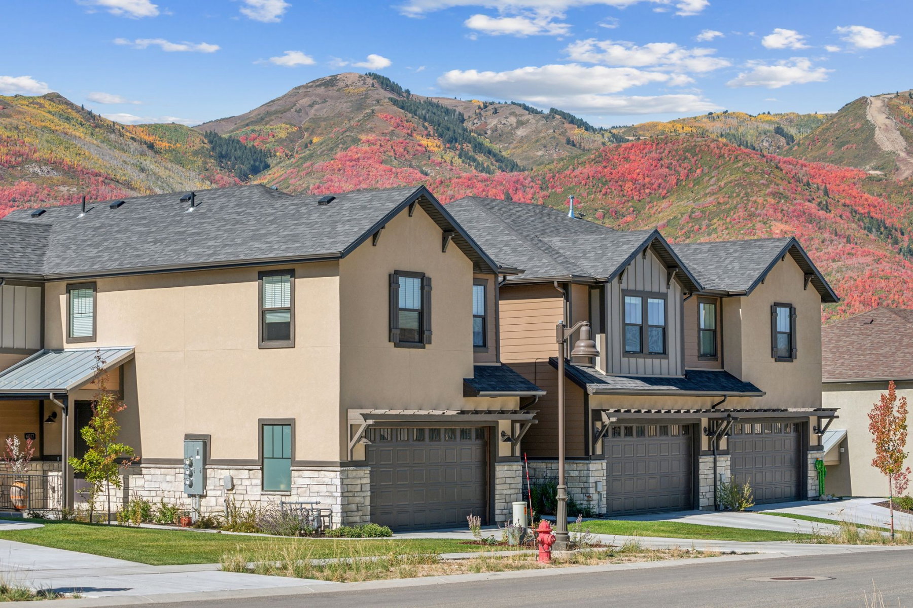 Townhome exterior featuring earth-toned stucco and wood siding, stone accents, attached garages with pergolas, and a shingled roofline