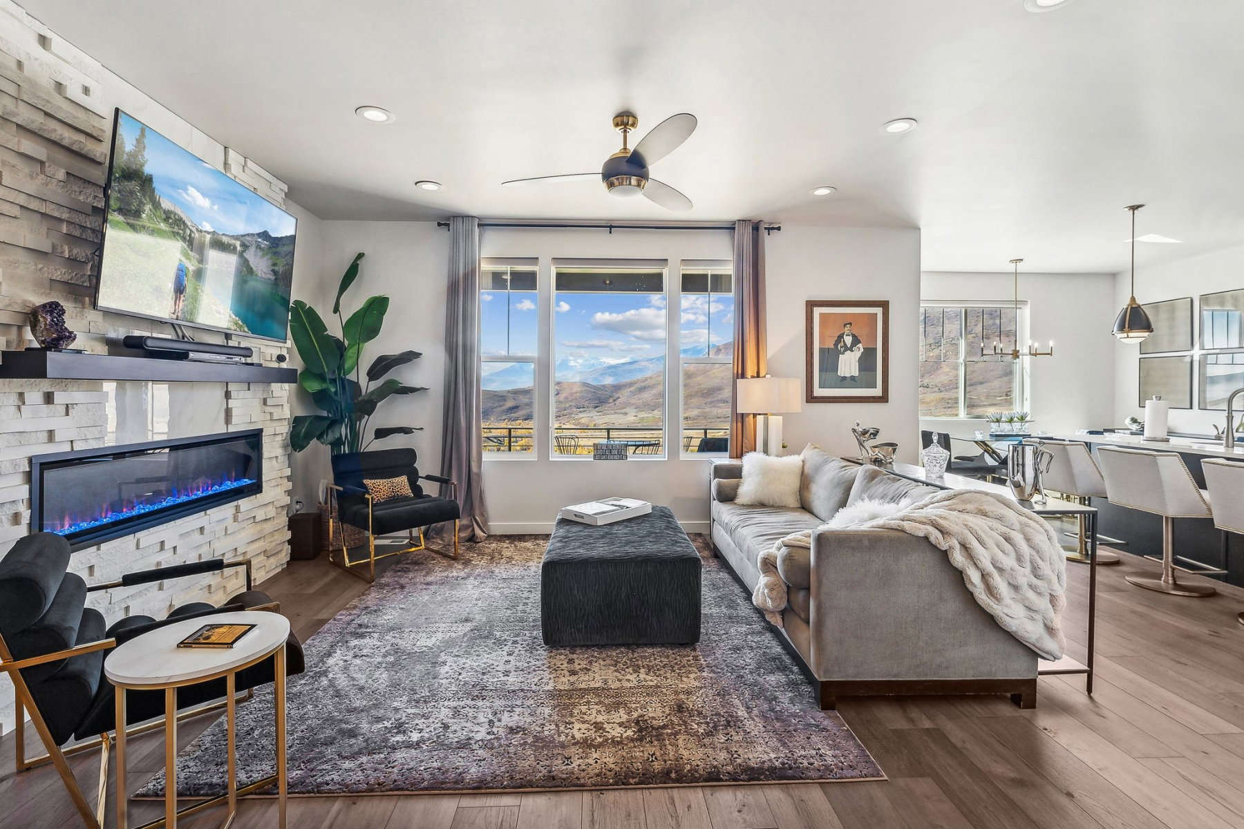 Spacious living area featuring a stacked stone fireplace with a built-in electric insert, wood-finish flooring, a ceiling fan, and large windows revealing a mountain landscape
