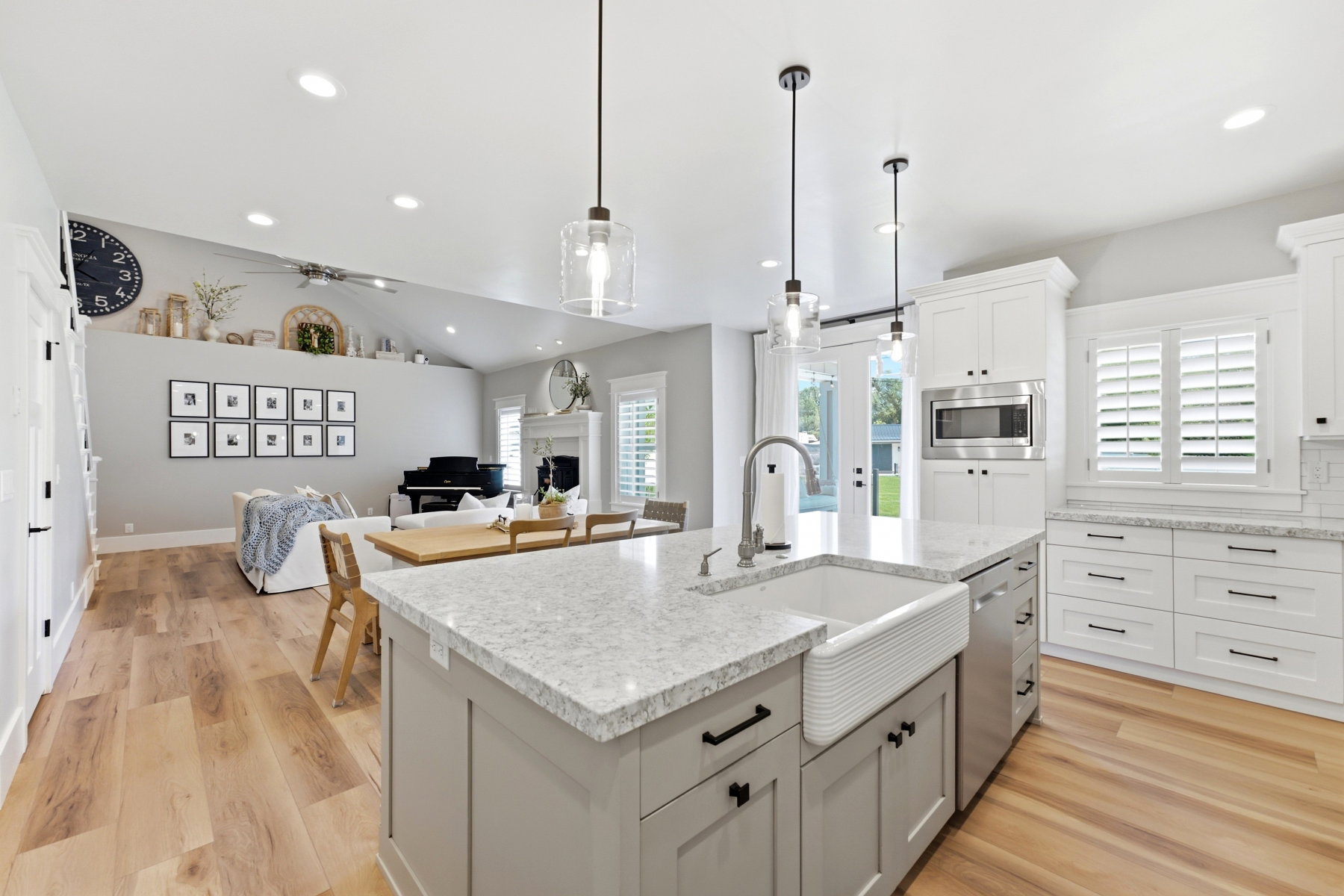 Open-concept kitchen featuring an oversized island with a farm sink, light-toned wood-finish flooring, white shaker cabinetry, stainless steel appliances, and plantation shutters