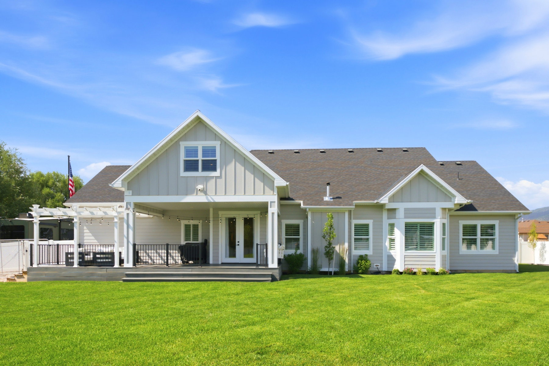 Expansive green lawn with a contemporary light gray siding exterior, featuring a covered patio, a separate deck with a white pergola, and multiple windows with white trim