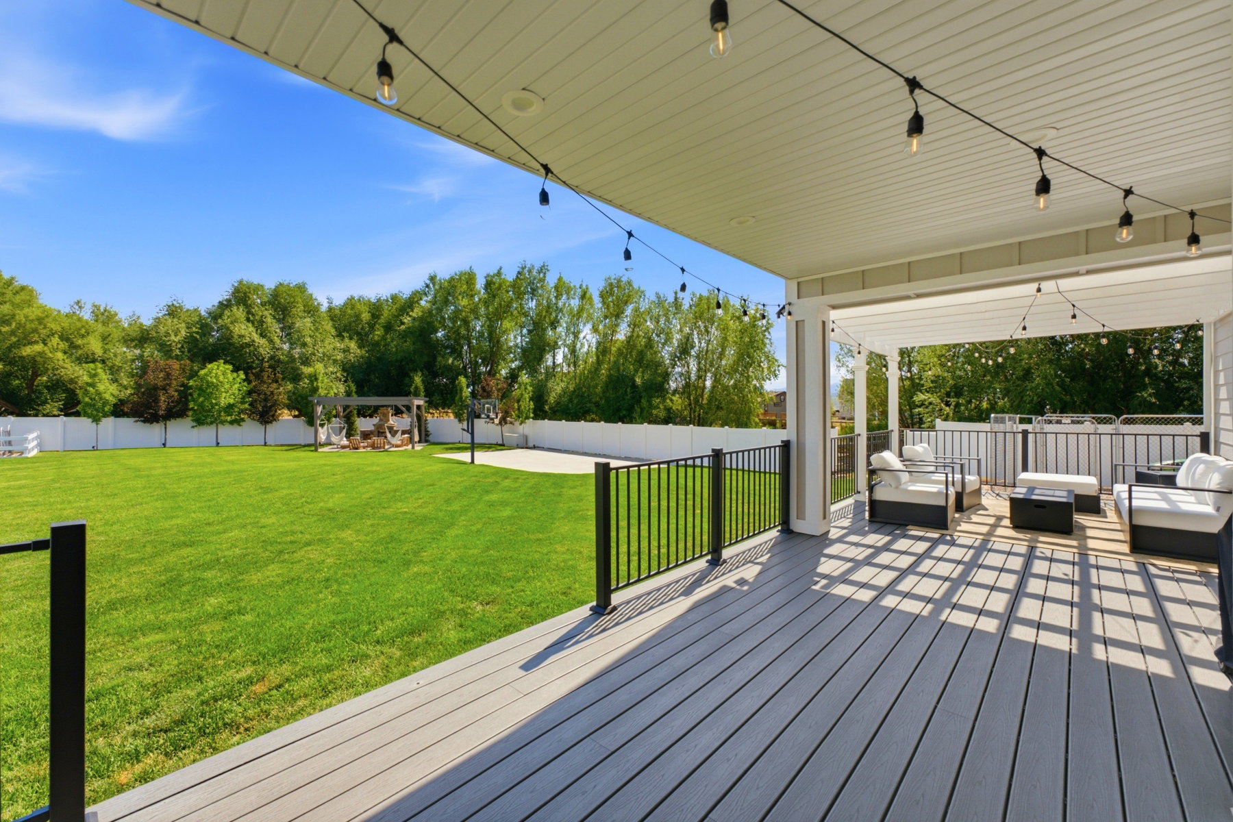 Expansive backyard featuring a covered composite deck, string lighting, a white privacy fence, and a separate paved patio area with a pergola