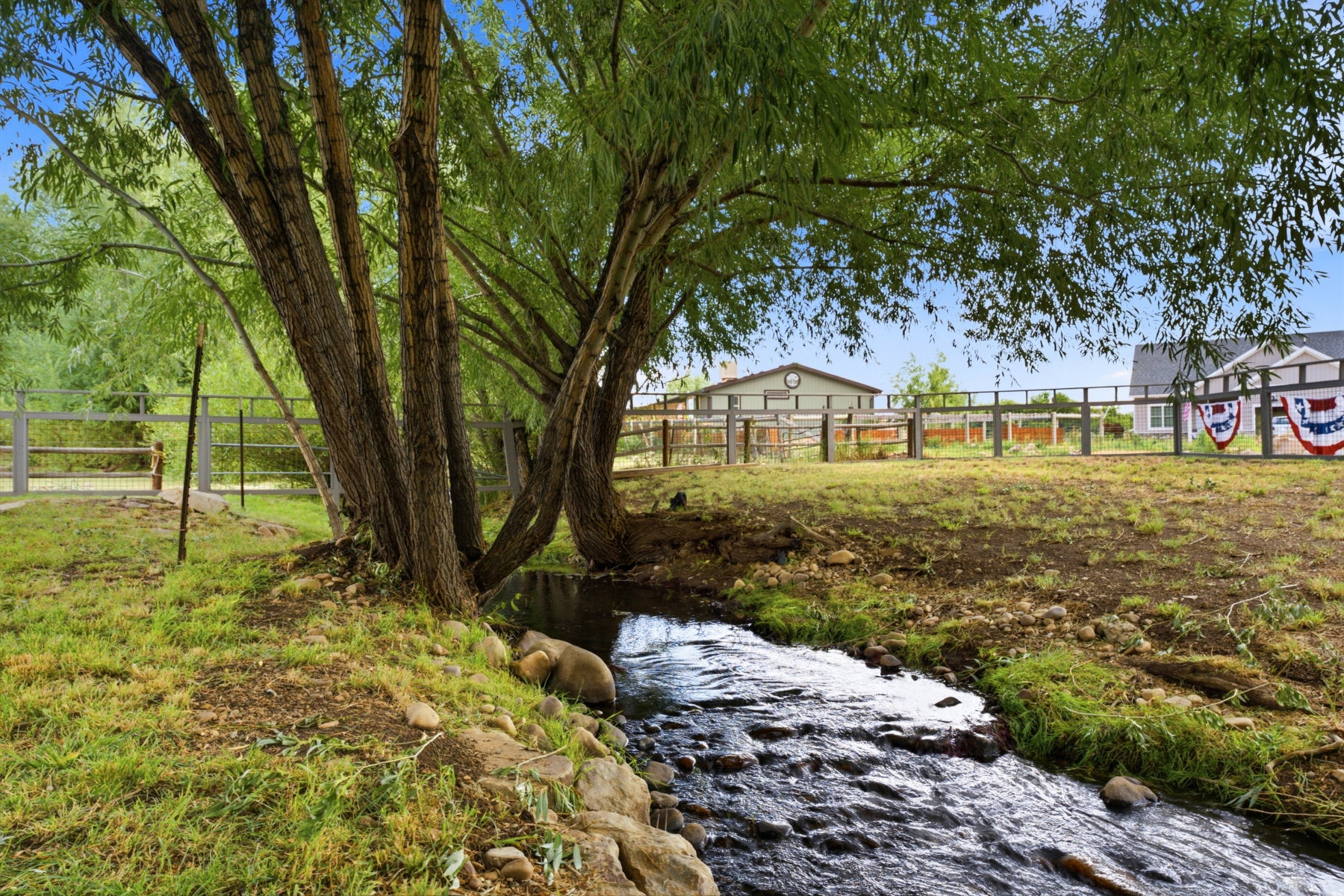 Natural stream feature with rock-lined banks, bordered by mature trees and a fenced perimeter
