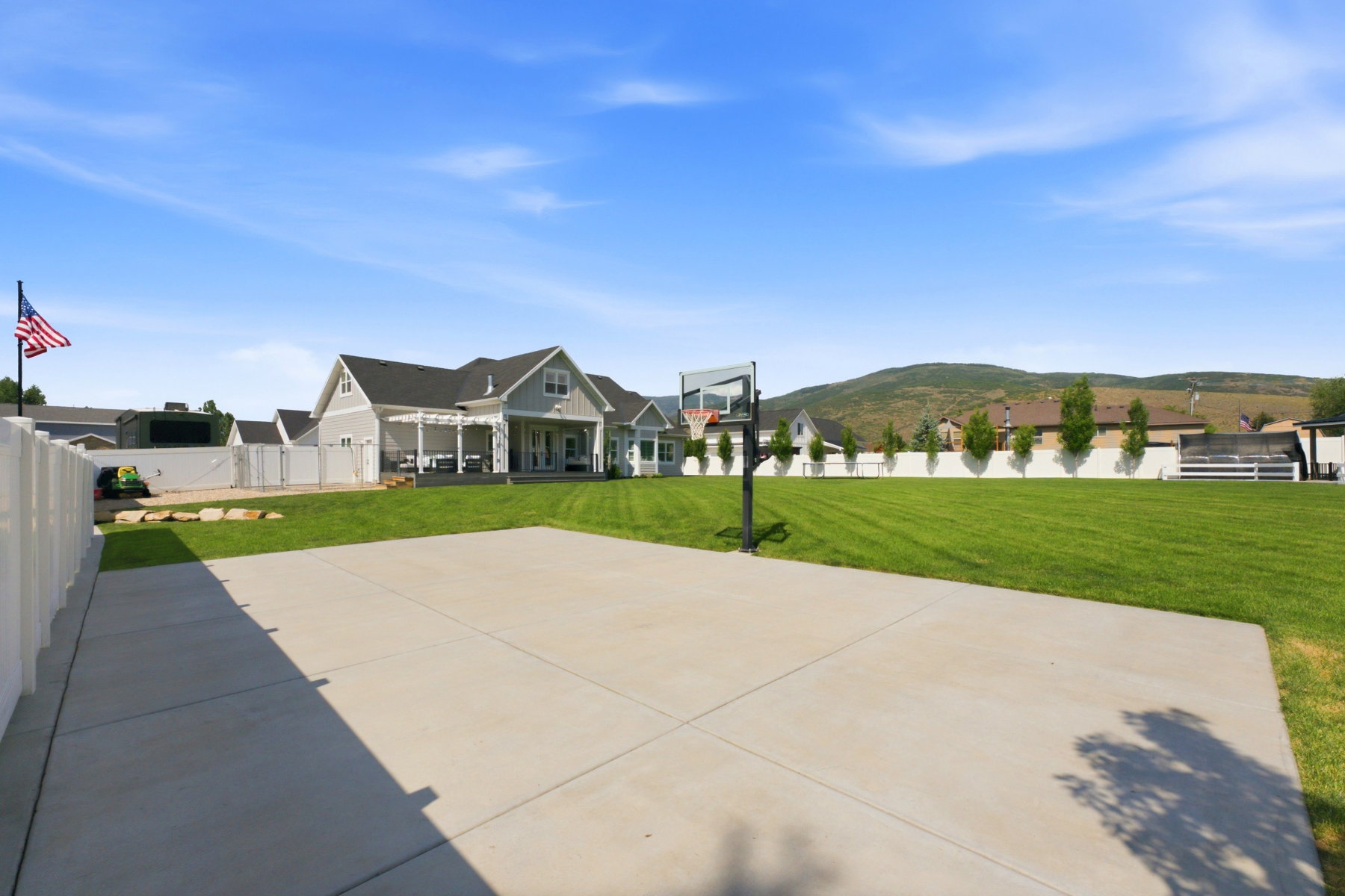 Expansive concrete patio with an integrated basketball hoop, bordered by a large manicured lawn and a white privacy fence