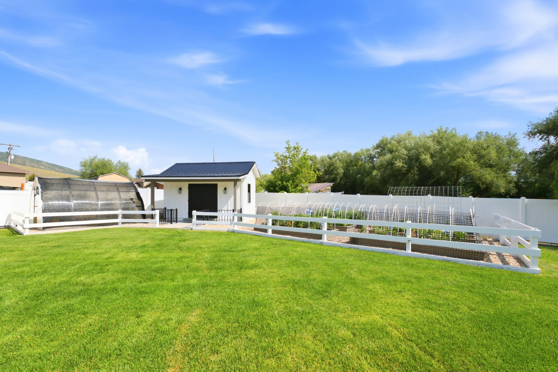 Spacious green lawn featuring a white privacy fence, a dedicated gardening area with raised beds, a white outbuilding with a dark metal roof, and an additional covered structure