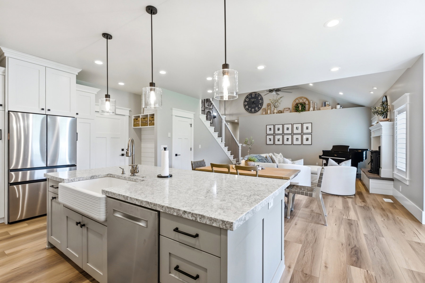Open-concept living area featuring a kitchen island with a farmhouse sink and light gray cabinetry, stainless steel appliances, wood-finish flooring, recessed lighting, and a white fireplace with a mantel