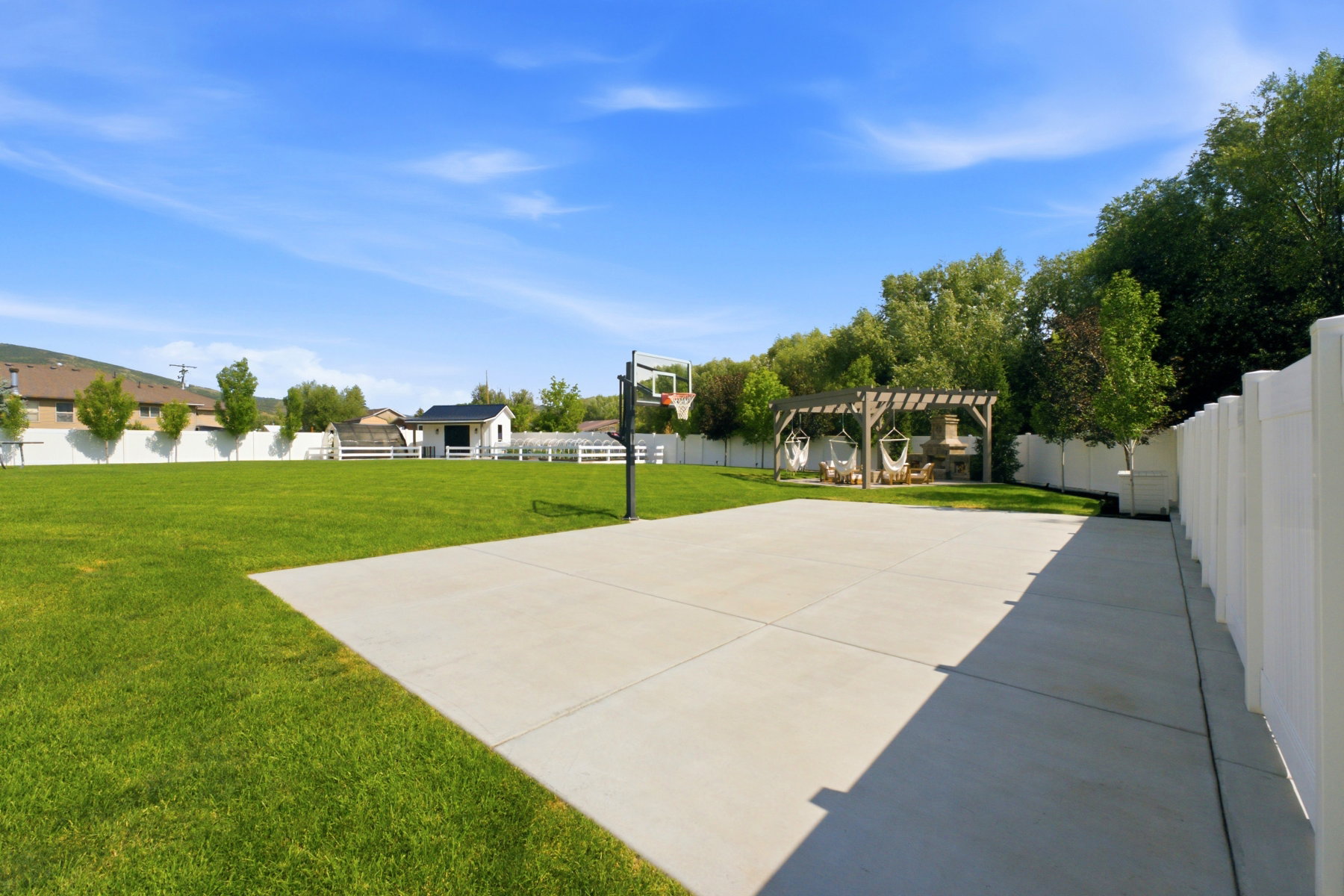 Expansive outdoor space featuring a concrete pad with a basketball hoop, a pergola with an outdoor fireplace, a manicured lawn, and white privacy fencing
