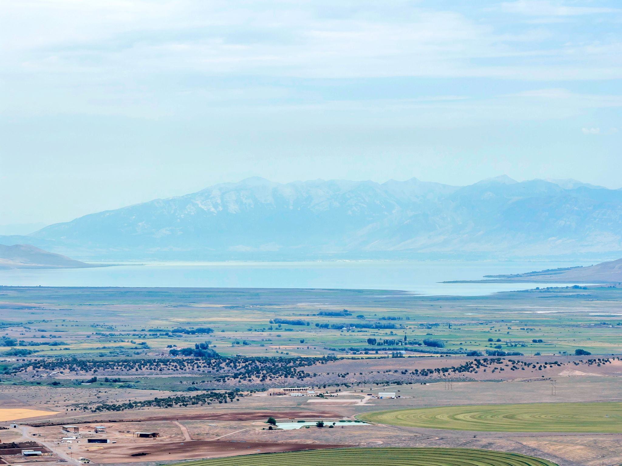 Expansive landscape featuring distant mountains, a large body of water, broad fields, and scattered structures