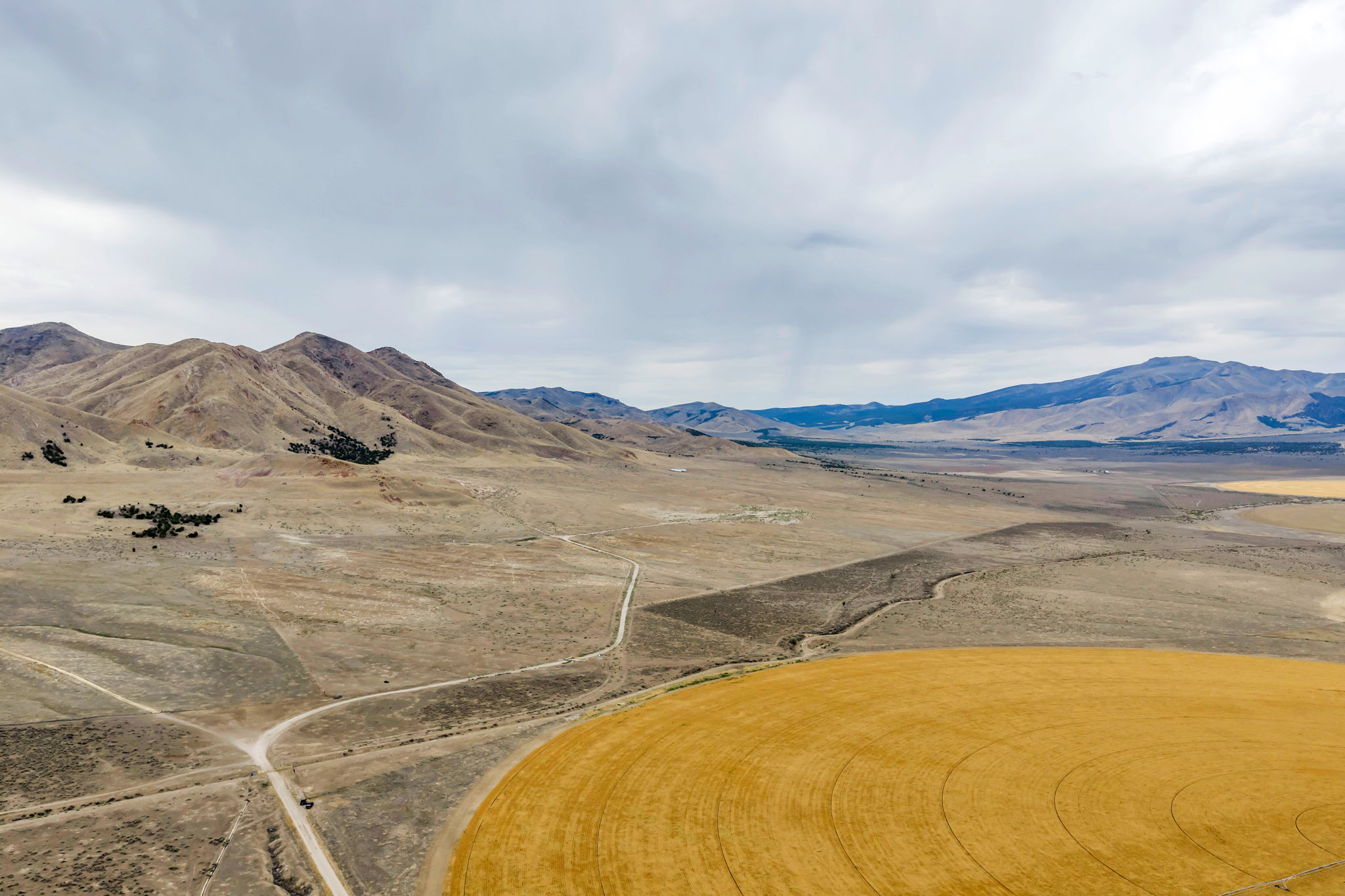 Expansive rural landscape featuring a cultivated field with circular patterns, unpaved roads, and distant mountains