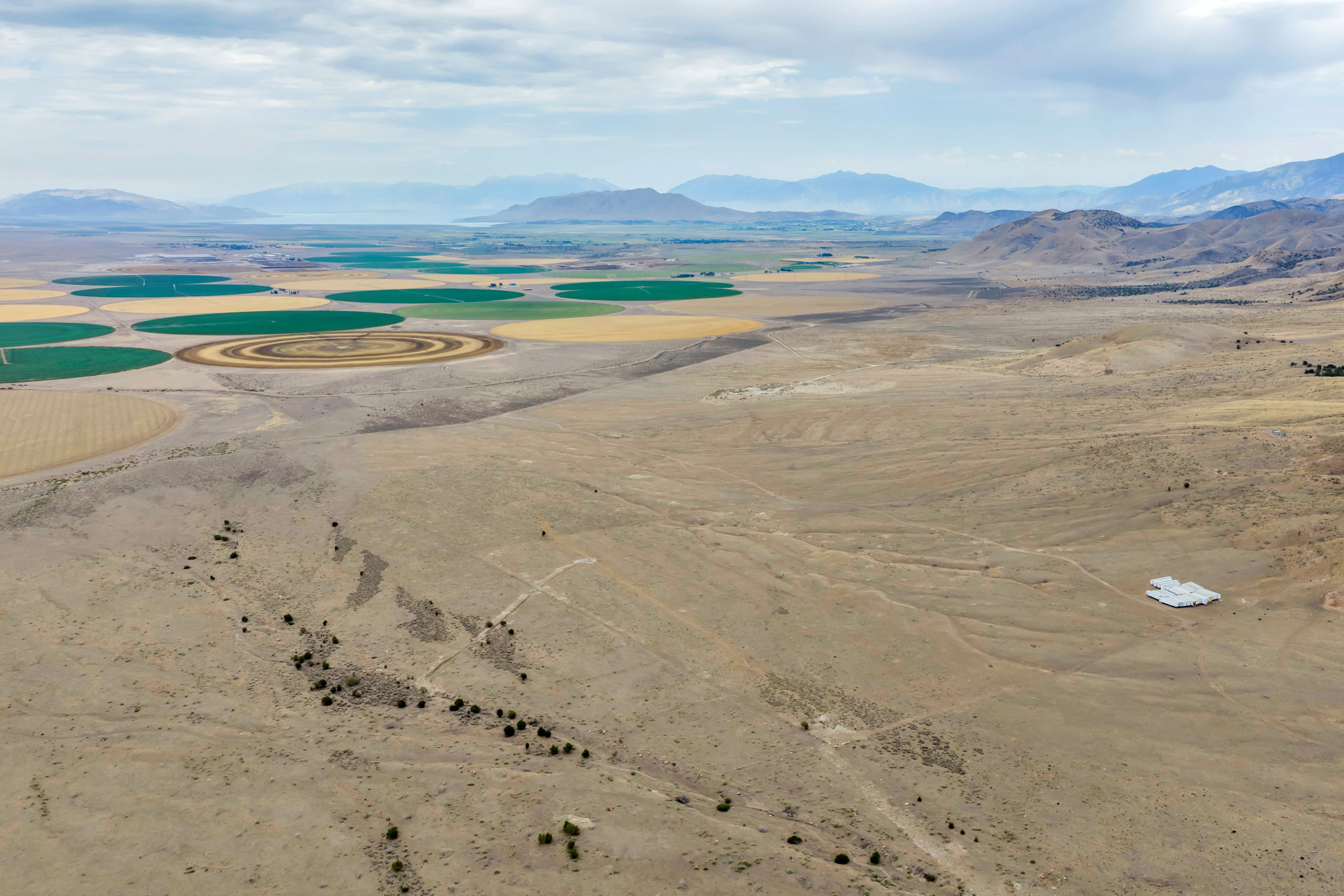Expansive arid land parcel featuring distant circular agricultural fields, scattered low-lying vegetation, a prominent white-roofed structure, and distant mountain ranges