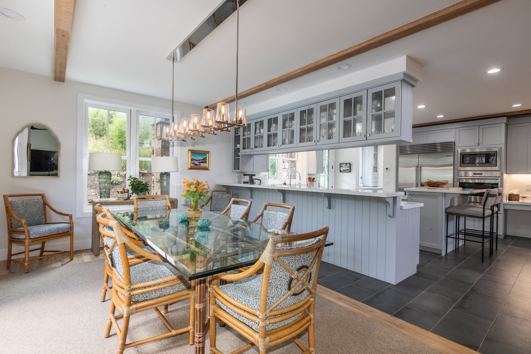 Open concept dining area featuring a linear chandelier, exposed wood beams, and a window framing outdoor greenery