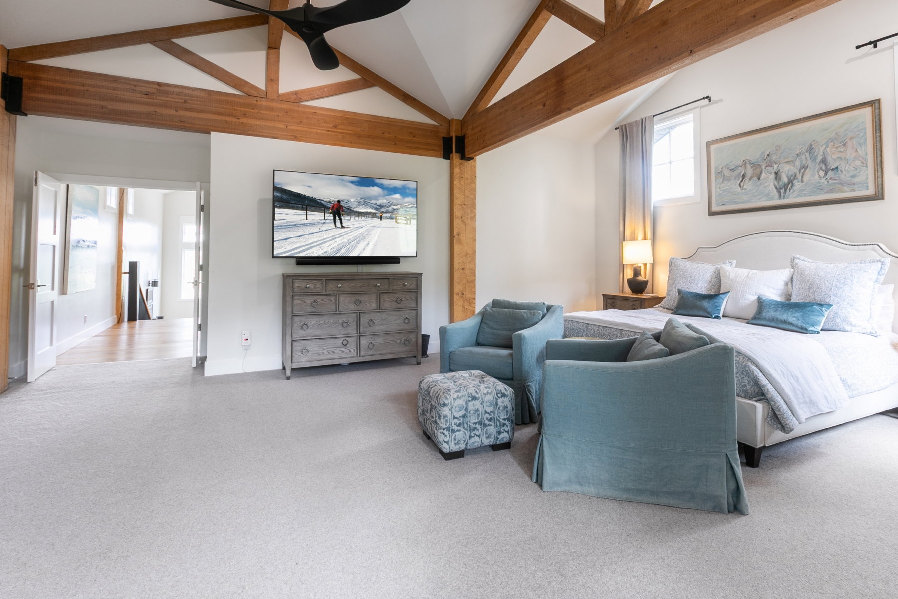 Vaulted ceiling with exposed wood beams, plush carpeting, white walls, and a window with a sheer curtain