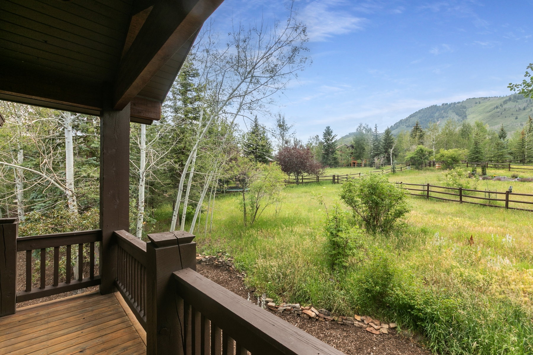 Expansive outdoor deck with natural wood flooring and railing, offering a sweeping landscape with mature trees, verdant fields, and mountain terrain