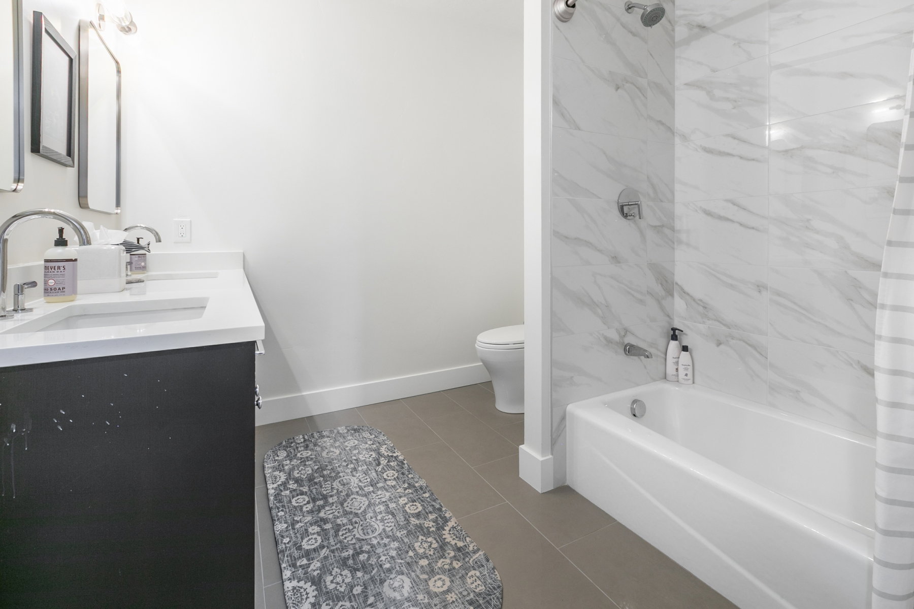 Bathroom featuring a dual-sink vanity with white countertops, a deep soaking tub, a tiled shower surround with a rainfall showerhead, and neutral floor tiling