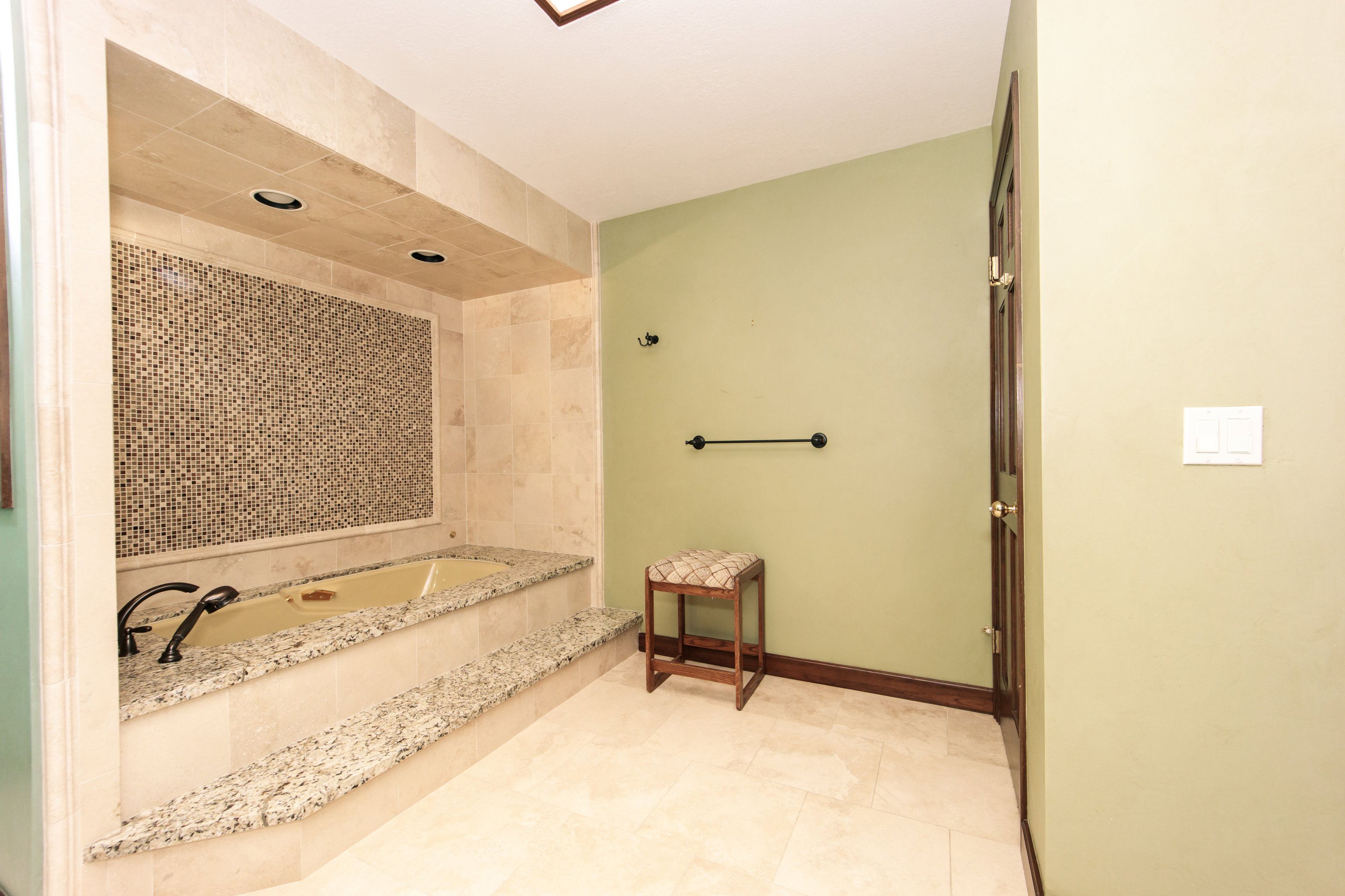 Recessed soaking tub with surrounding stone tile, mosaic-tiled accent wall, and granite countertop