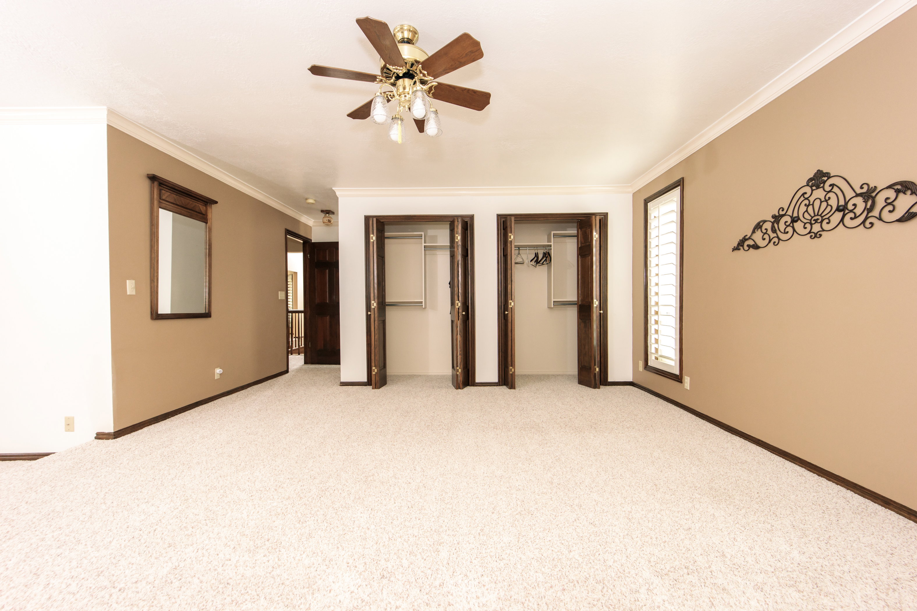 Spacious room featuring cream-colored carpeting, two double-door closets, crown molding, a ceiling fan with integrated lighting, and a window with white louvered shutters