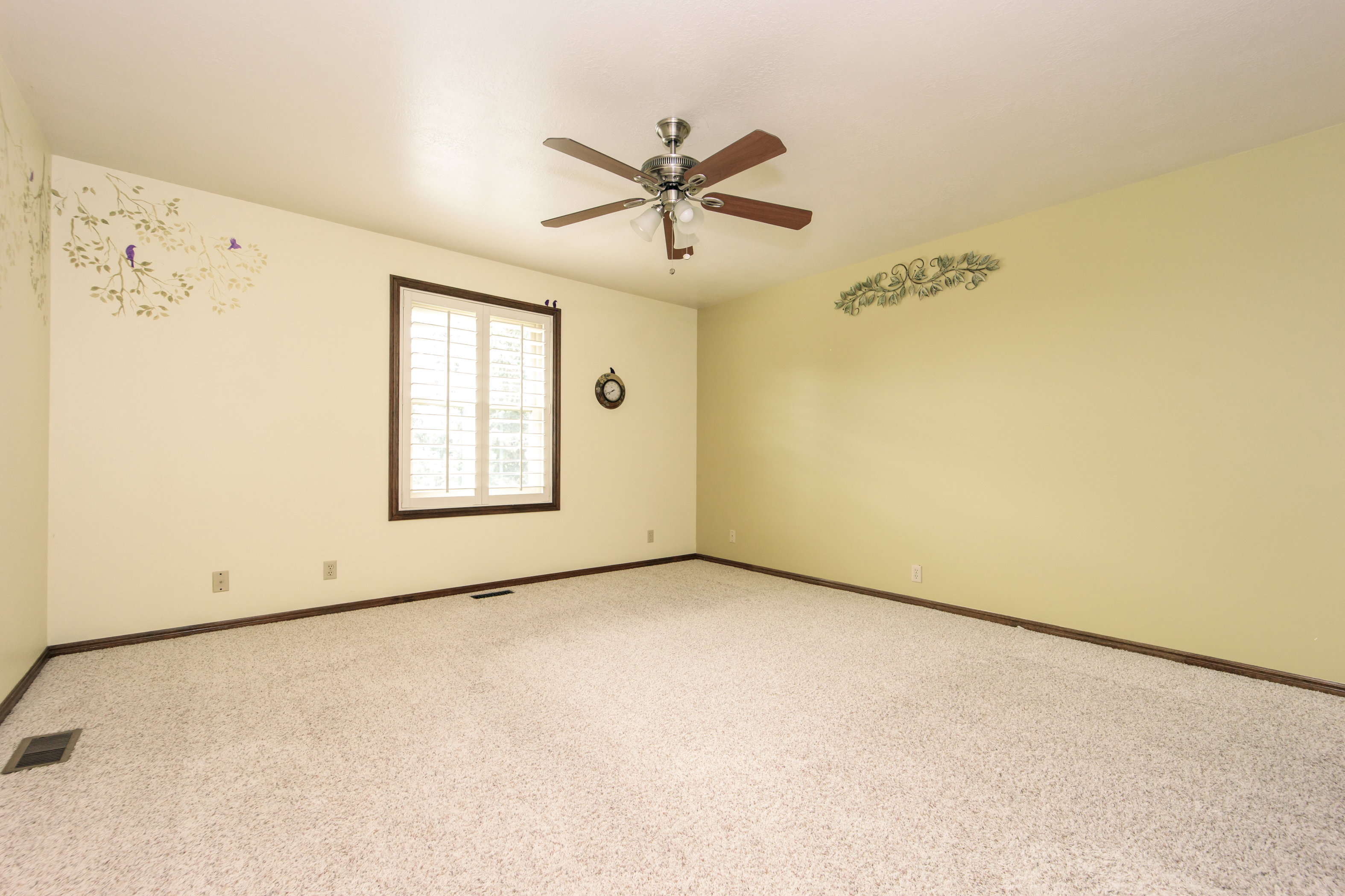 Spacious room featuring light-colored carpeting, a ceiling fan with light fixture, and a window with white louvered shutters and dark wood trim