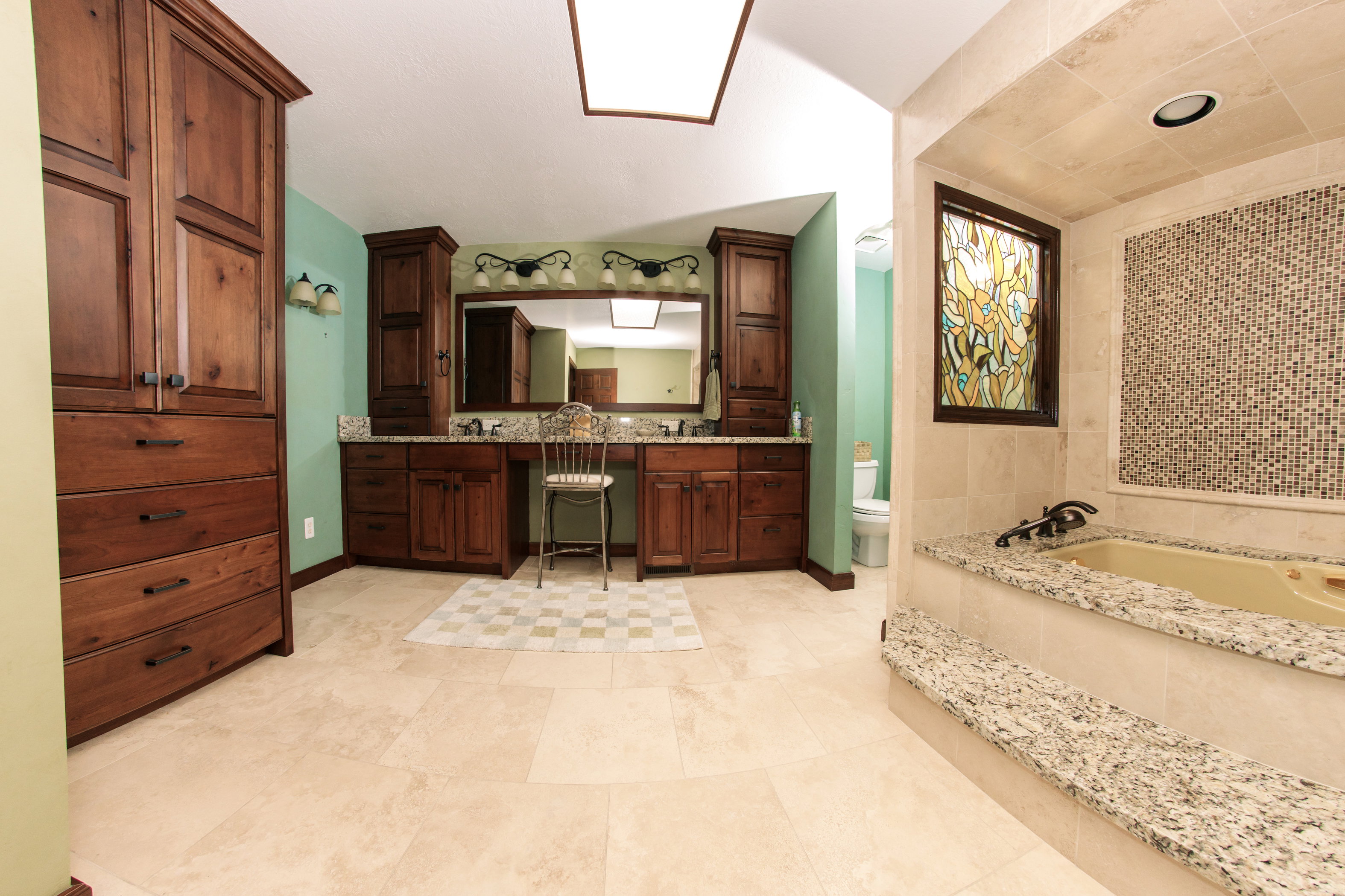 Spacious bathroom featuring light-toned tile flooring, extensive wood cabinetry, and a double vanity with granite-finish countertops
