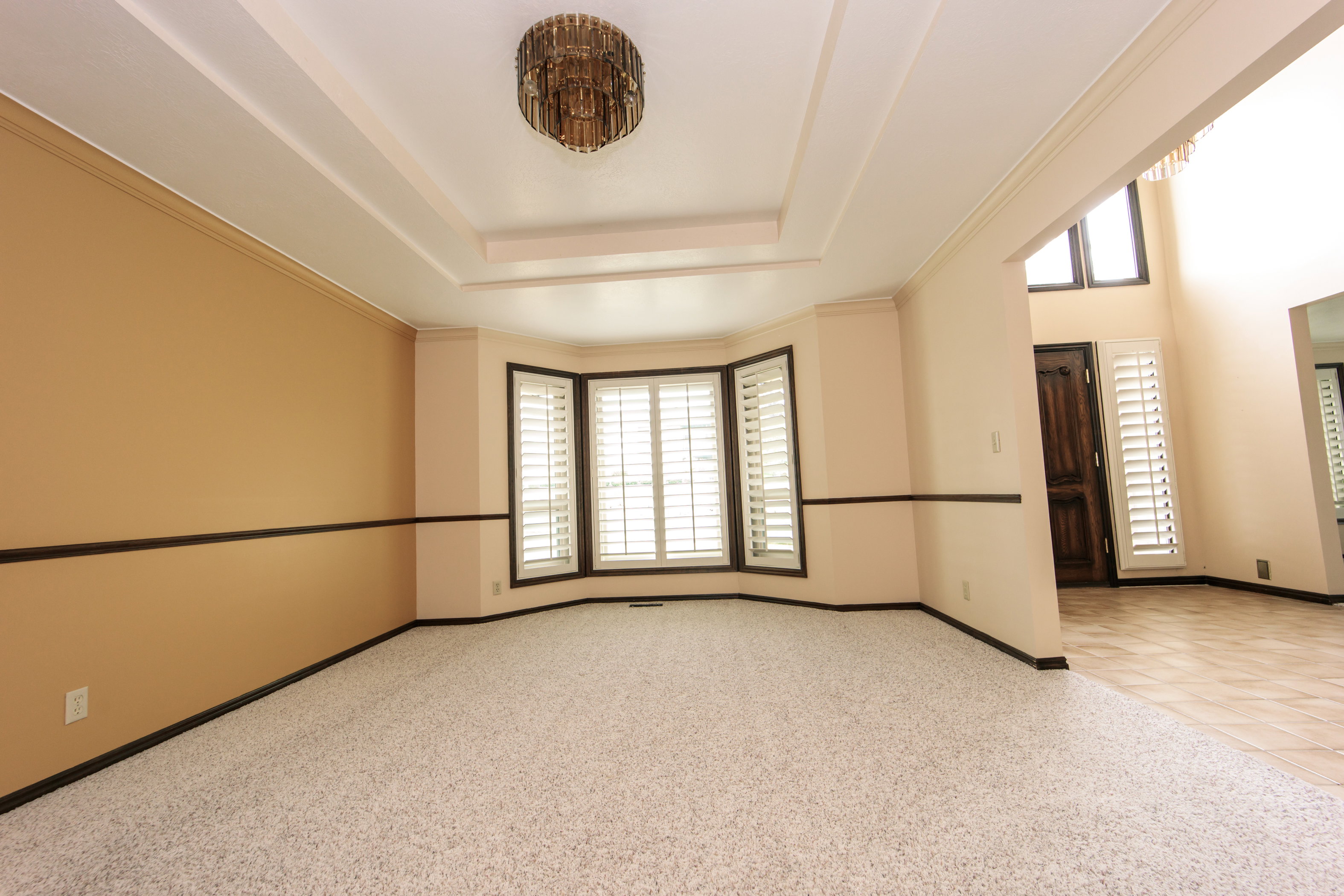 Recessed ceiling details with a central chandelier illuminate this room featuring a bay window with plantation shutters
