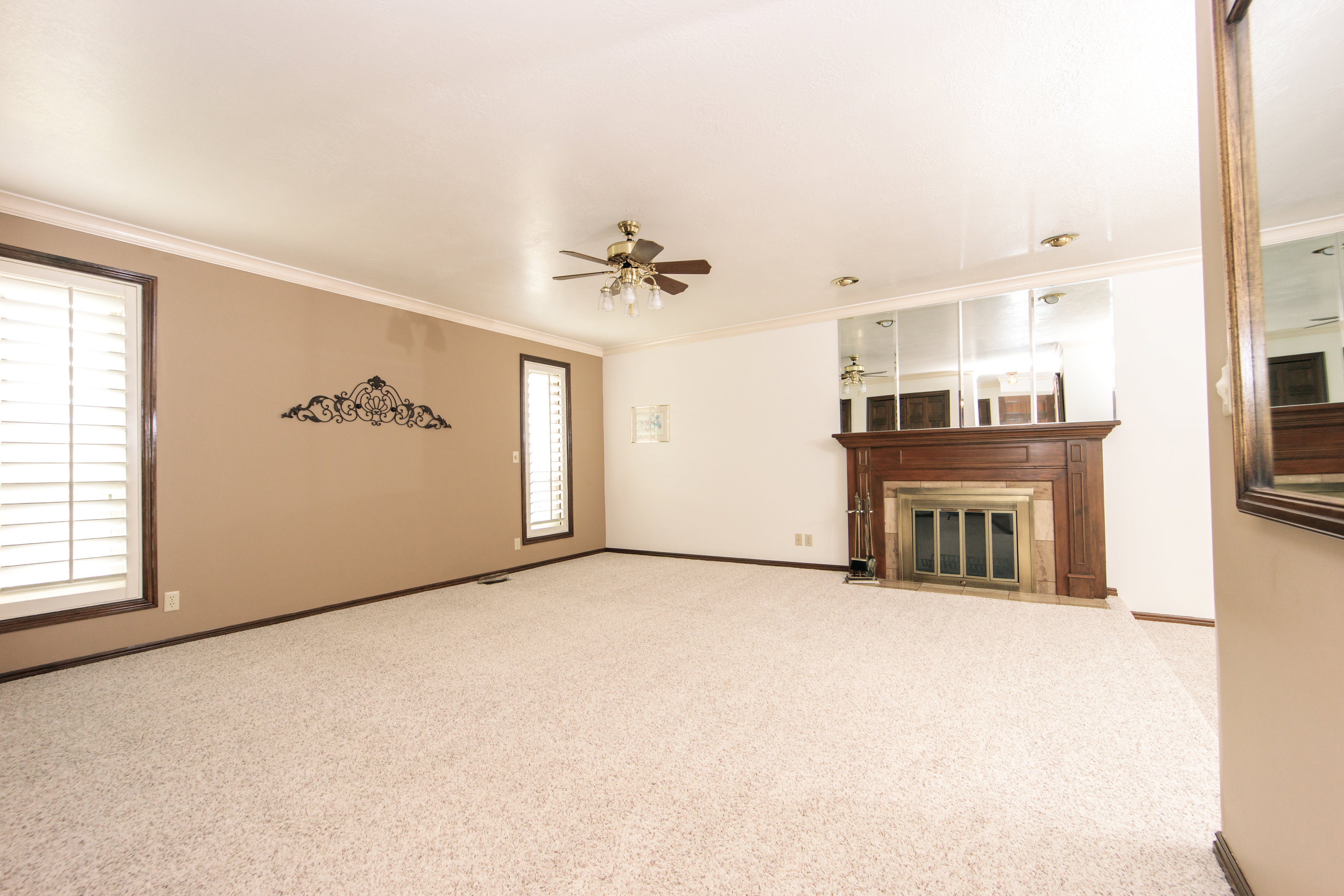 Spacious carpeted interior featuring a wood-mantled fireplace, mirrored accent wall, and ceiling fan with integrated lighting