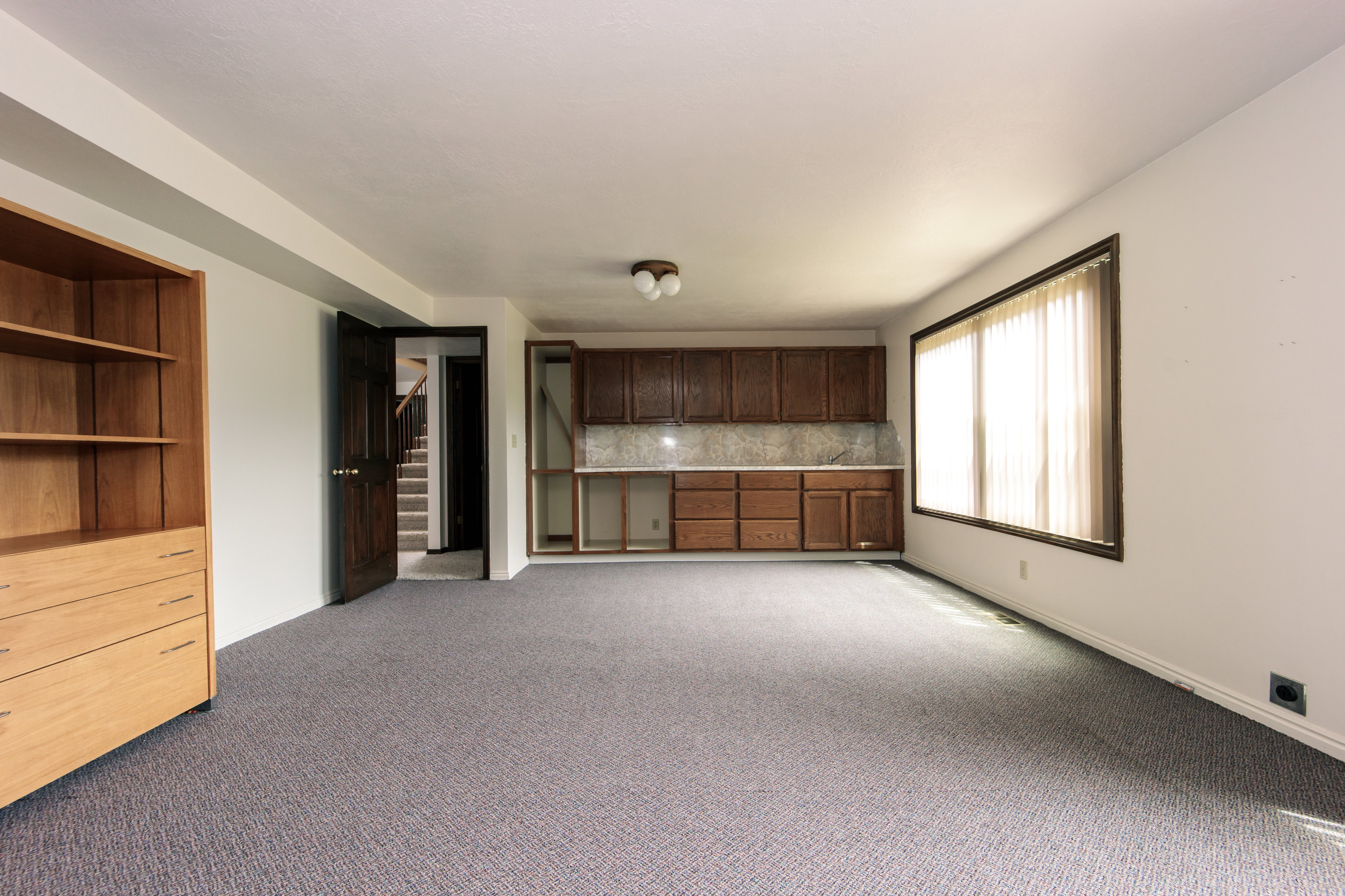 Expansive room featuring wood-finish built-in shelving, dark wood-finish cabinetry with light-toned backsplash, a wide window, and neutral carpeting