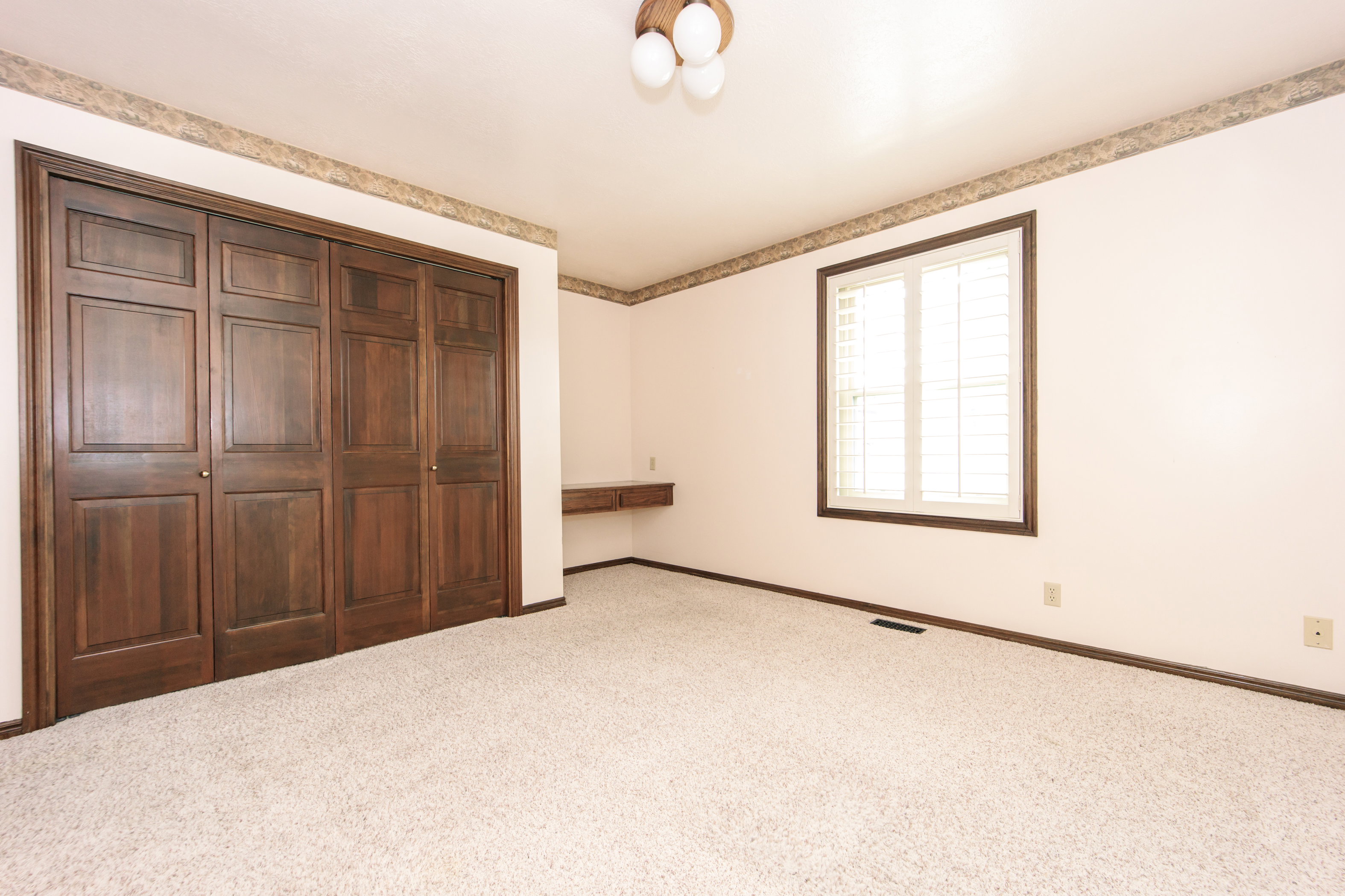 Spacious room featuring light-colored carpeting, a large window with white plantation shutters and dark wood trim, and extensive dark wood bi-fold closet doors