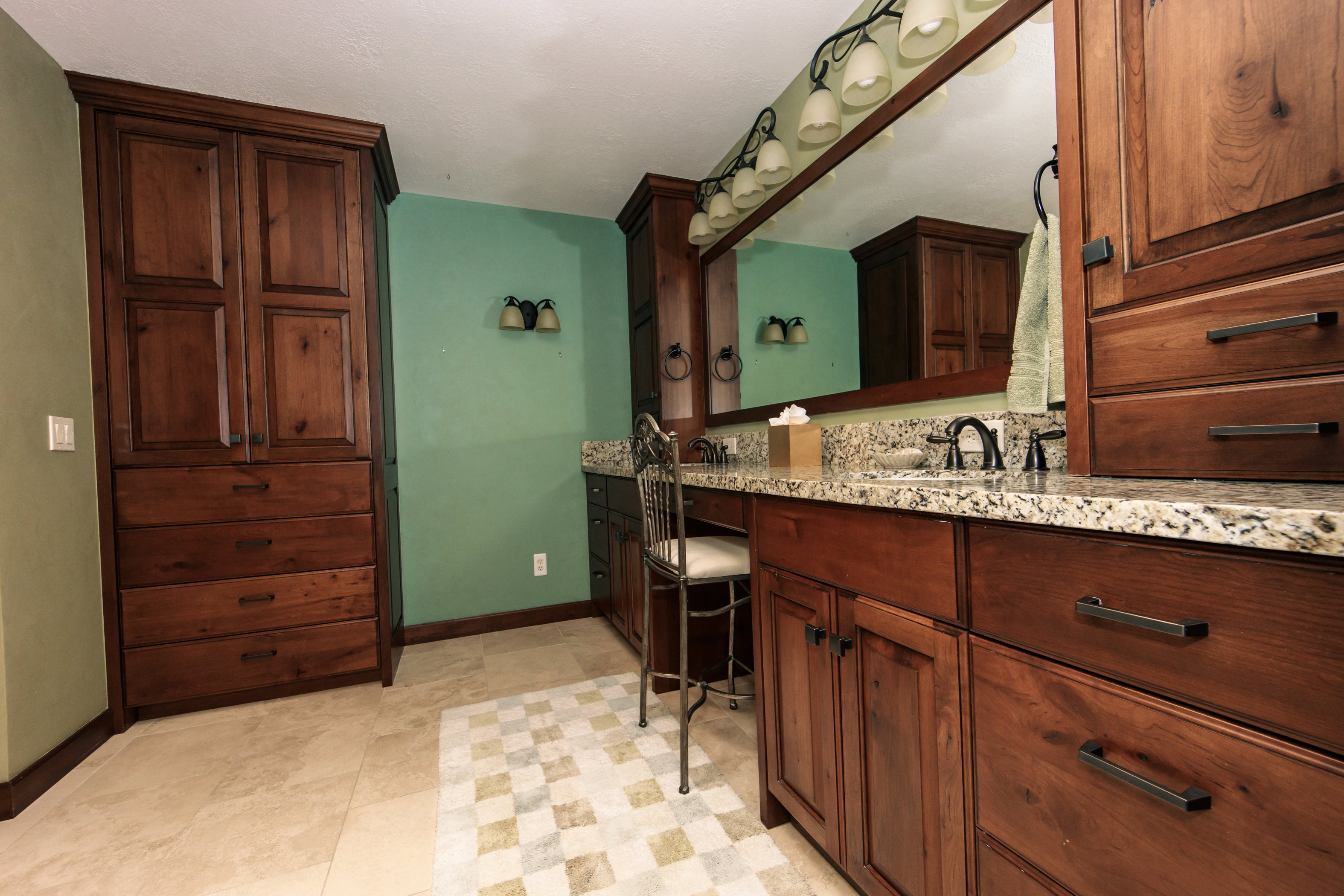Bathroom vanity featuring dual sinks with dark bronze fixtures, granite countertop, expansive wall-mounted mirror with overhead lighting, and rich wood cabinetry