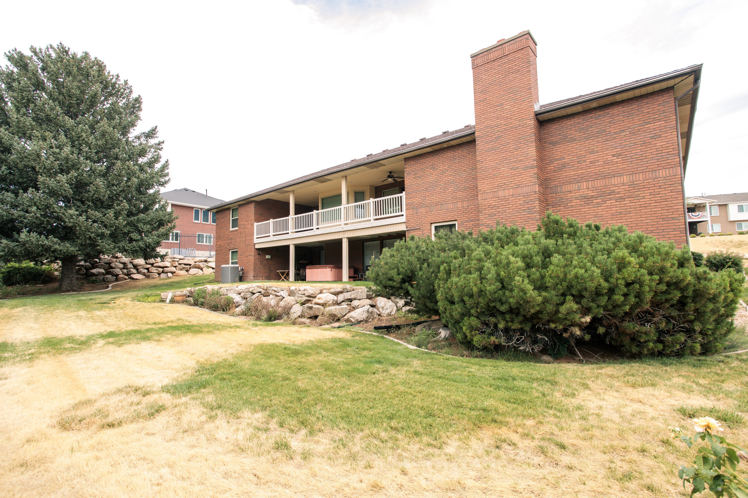 Brick exterior with a prominent chimney, upper-level balcony featuring white railings, lower-level patio area, and mature landscaping