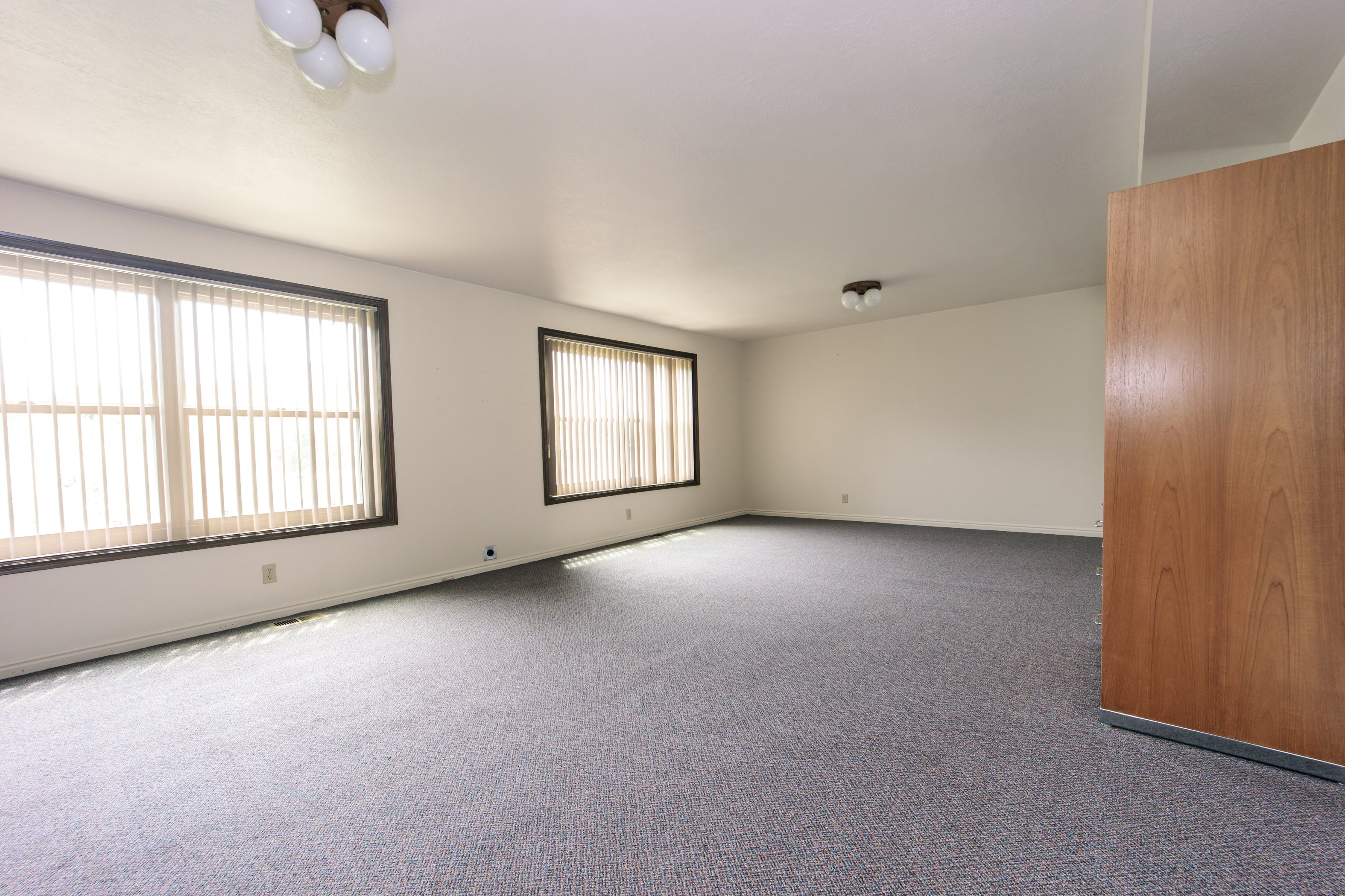 Spacious room featuring gray carpeting, multiple windows with vertical blinds, and a light wood-finish door