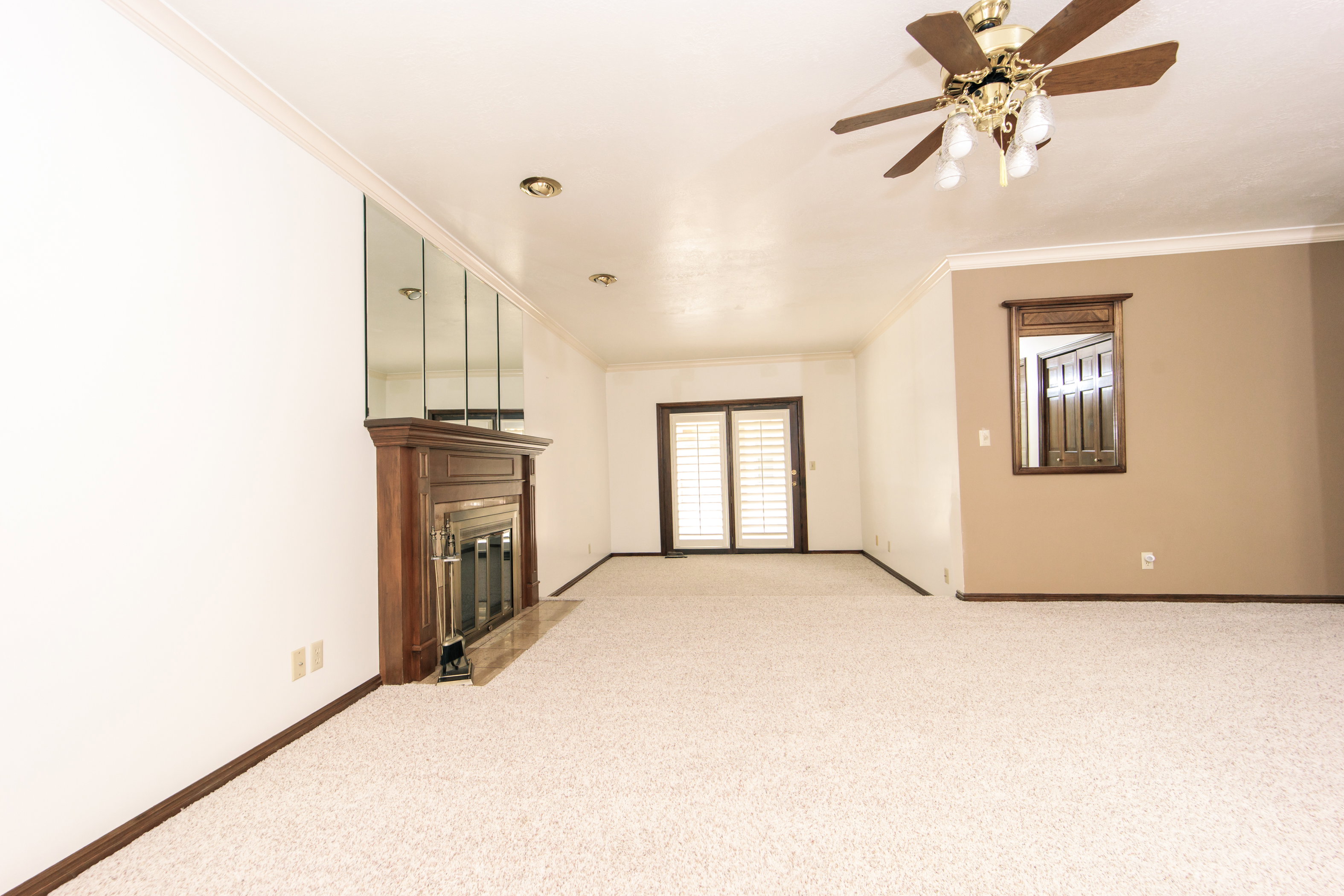Spacious room featuring light-toned carpet flooring, a wood-finish fireplace with a mirrored surround, recessed lighting, and a ceiling fan with integrated lighting